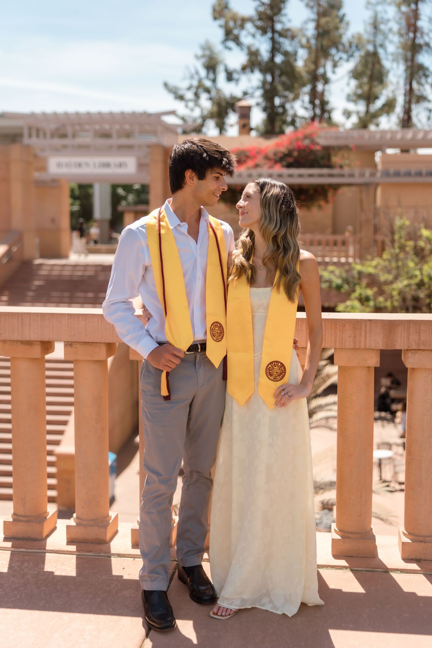 Two smiling college graduates in gold stoles share a joyful moment on a campus terrace.