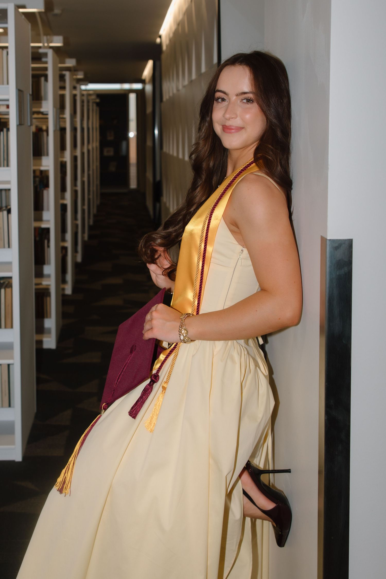 Young woman in cream dress with maroon and gold graduation stole holds cap in library hallway.