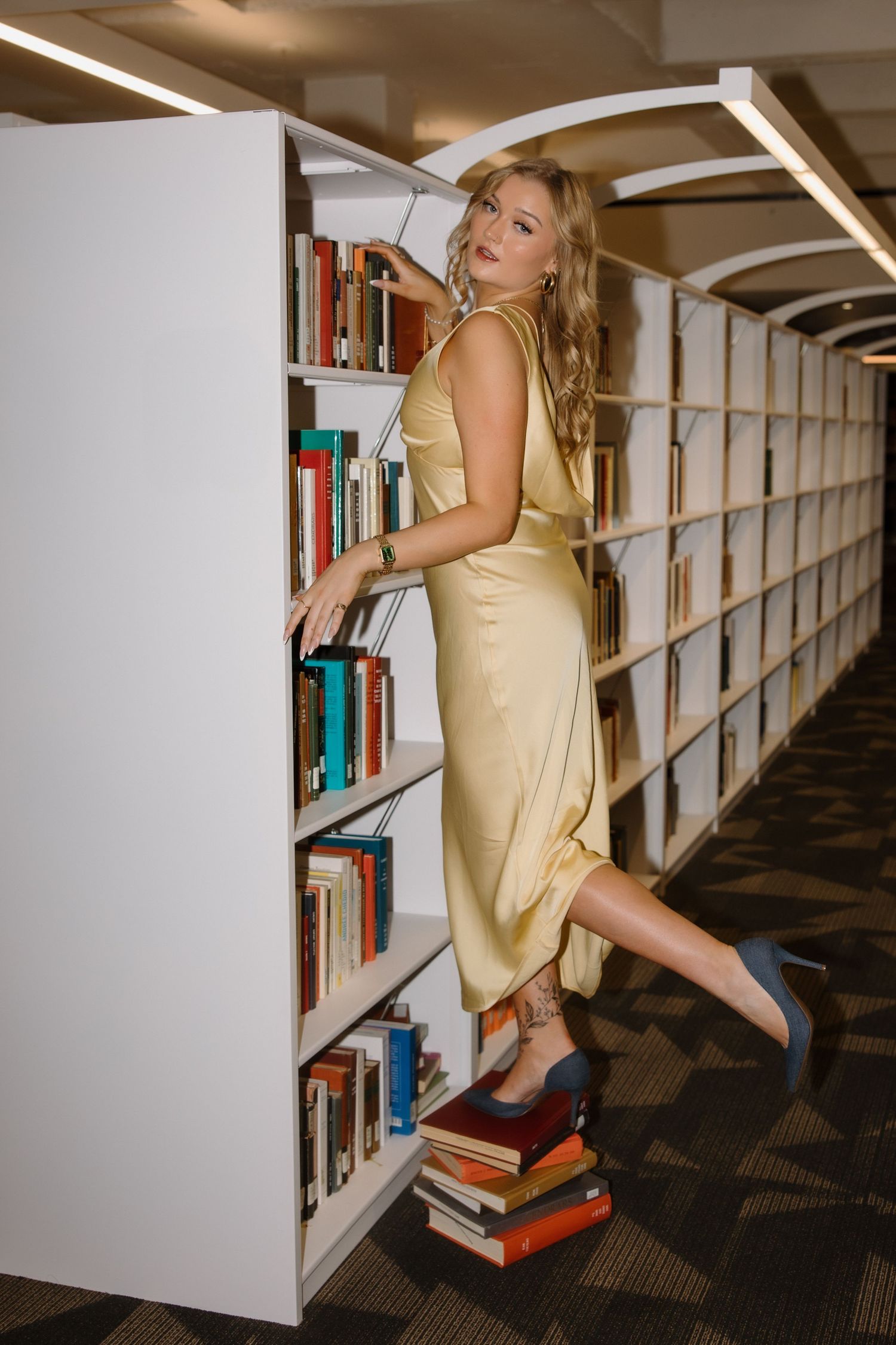Woman in gold dress standing on books reaching for shelf in modern library aisle with white bookcases.