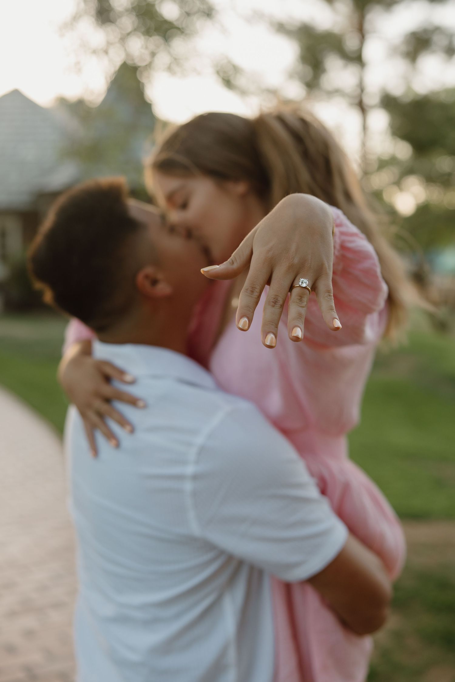 Couple sharing a romantic kiss during engagement, woman in pink dress showing off diamond ring while being lifted by man in white shirt.
