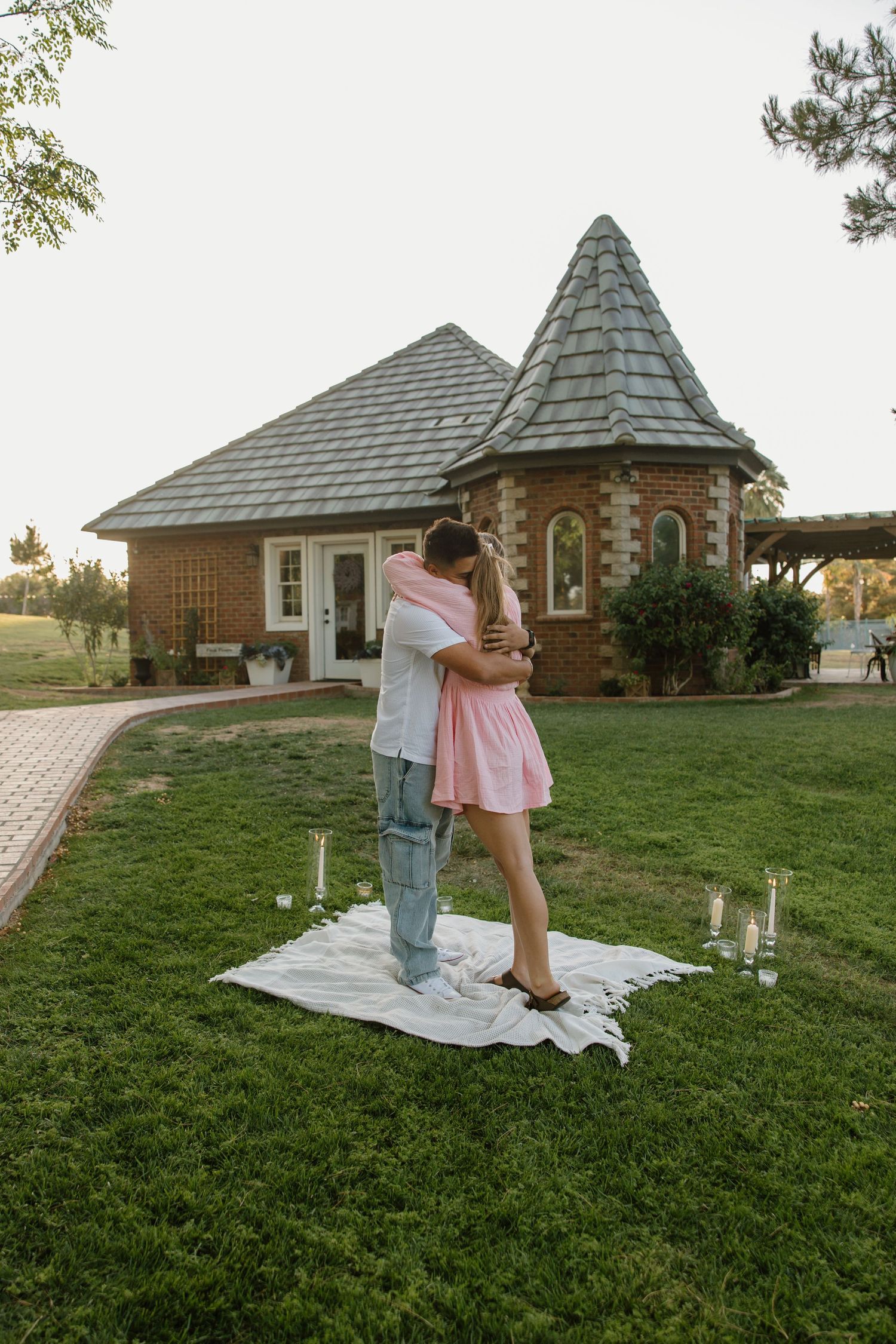 Couple embracing on a white blanket with candles during a romantic outdoor proposal in front of a charming brick building.