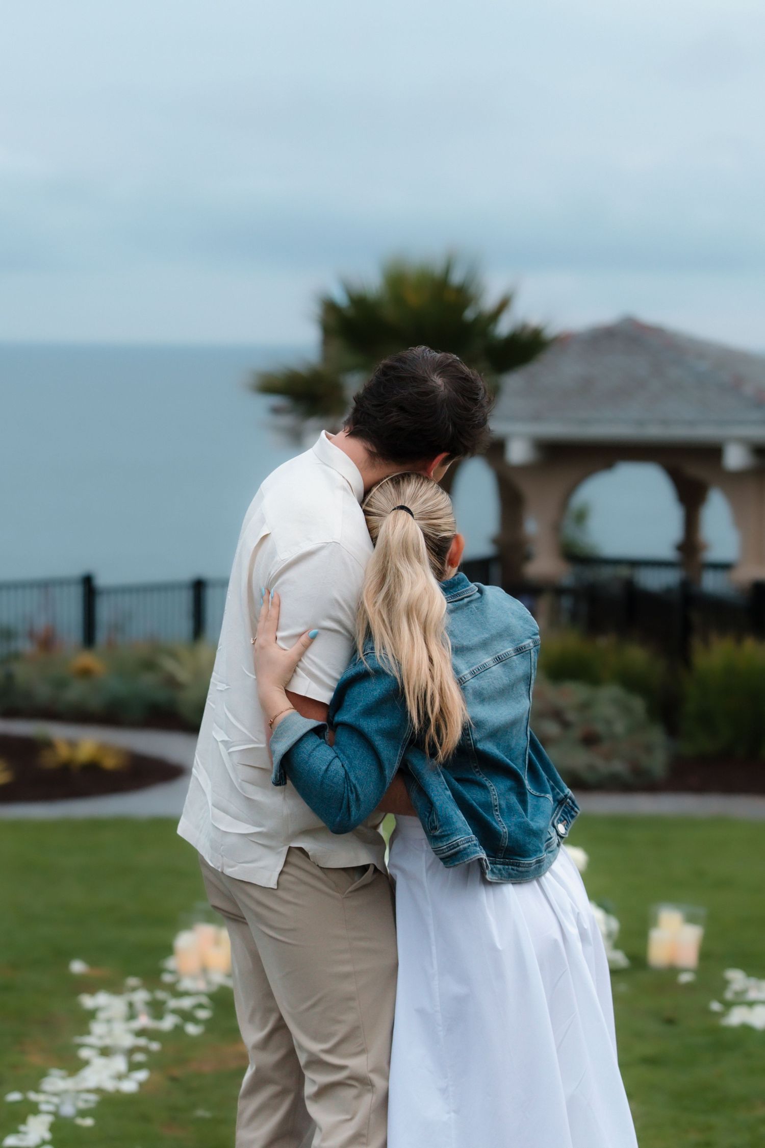 Newly engaged couple sharing tender embrace from behind near ocean overlook with white rose petal path.