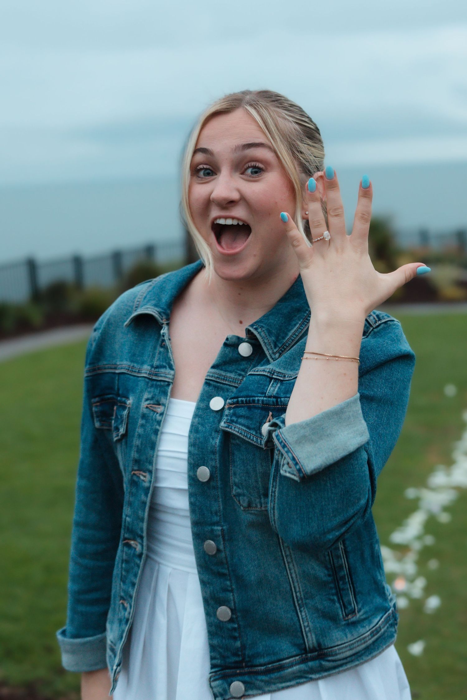 Excited blonde woman showing off new engagement ring with open mouth smile outdoors.