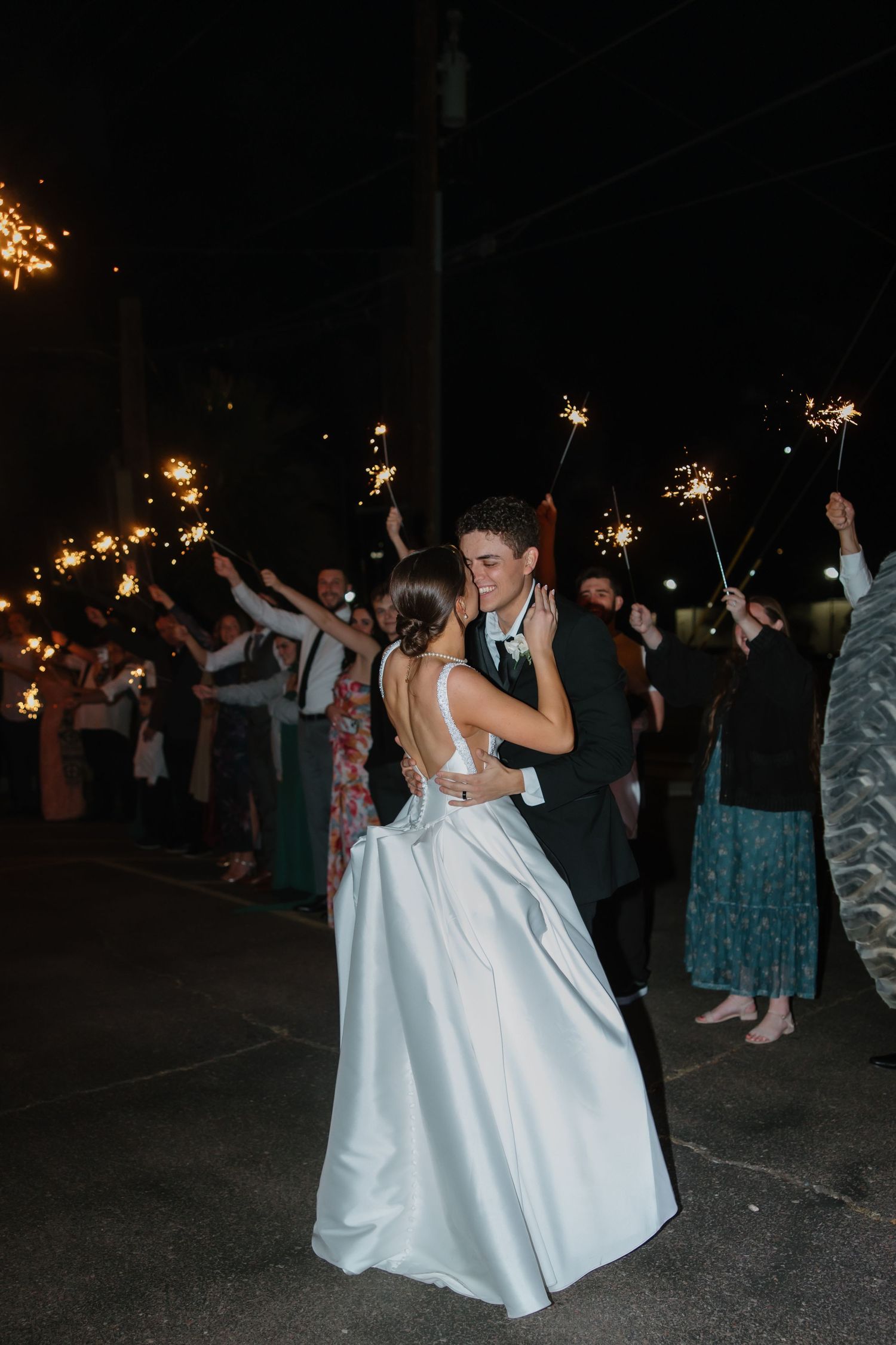 Bride in elegant white gown and groom dance romantically amid guests waving sparklers at night wedding exit.