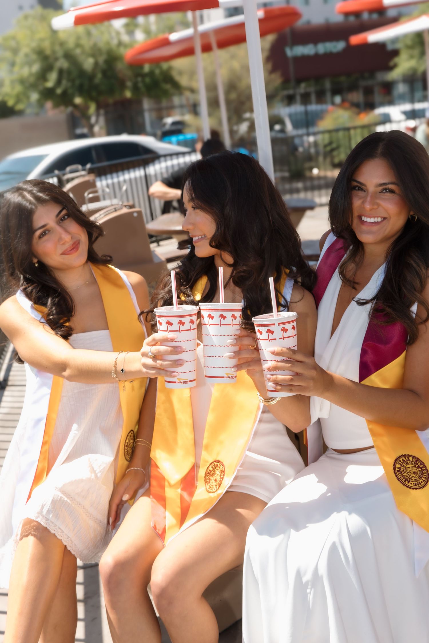 Three smiling graduates in white dresses and gold stoles toast with In-N-Out drinks at an outdoor table.