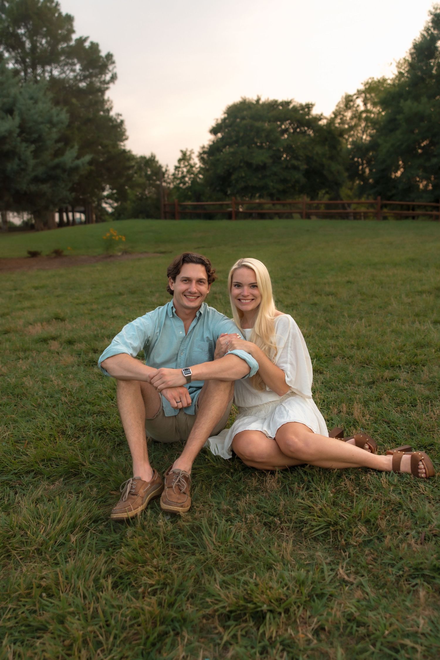 Happy couple sitting together on grass in park, smiling at camera during golden hour sunset.