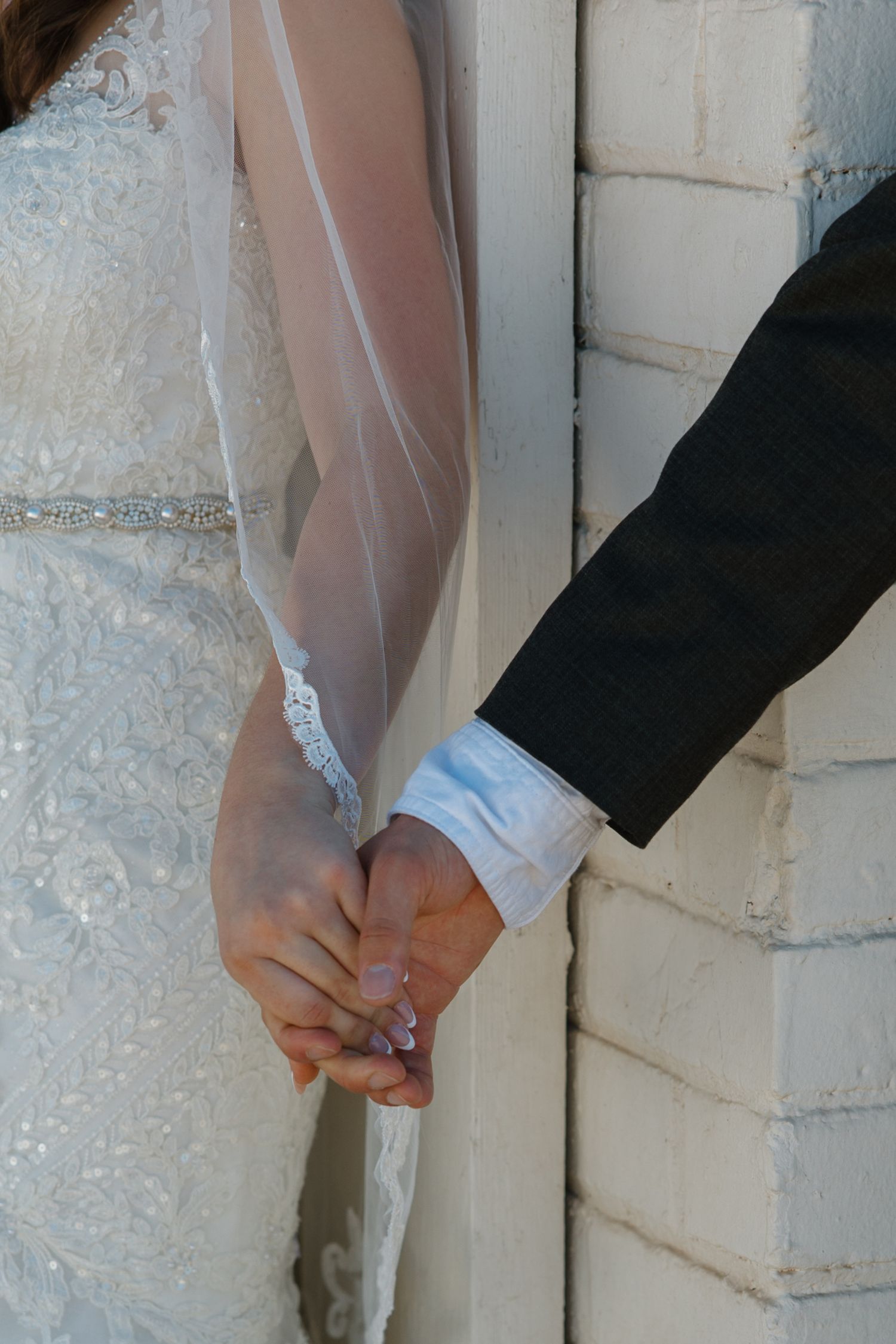 Bride and groom holding hands at wedding, bride in lace gown with veil, groom in black suit, against white brick wall.