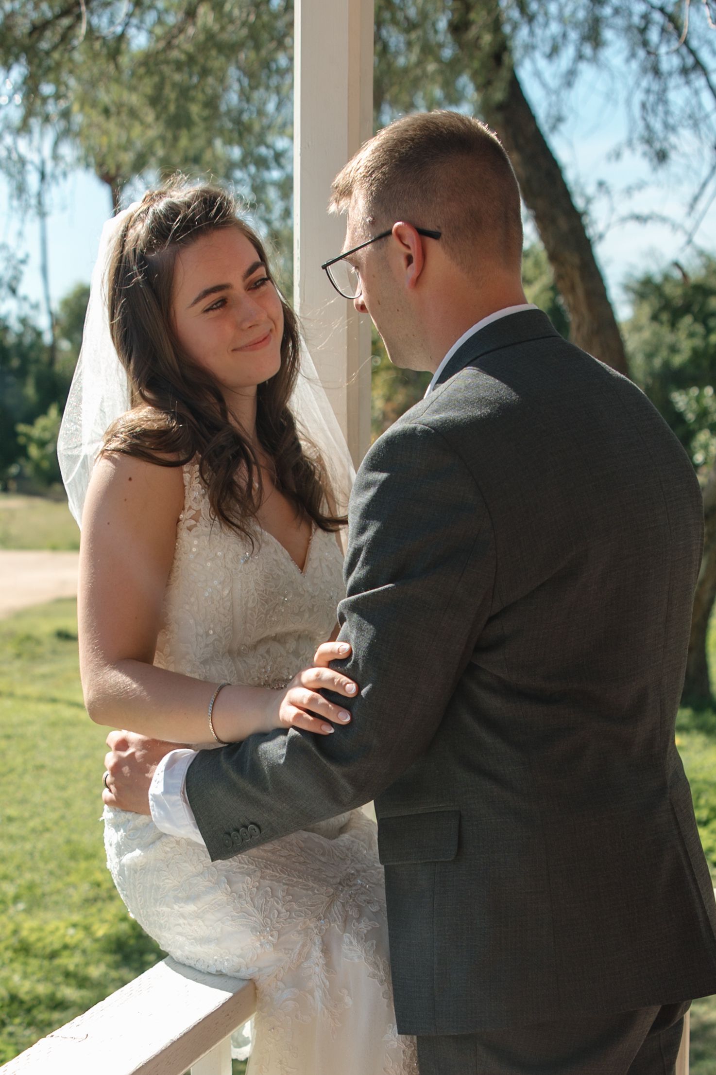 Smiling bride in lace gown holds groom's arm near white pillar during outdoor wedding portrait.