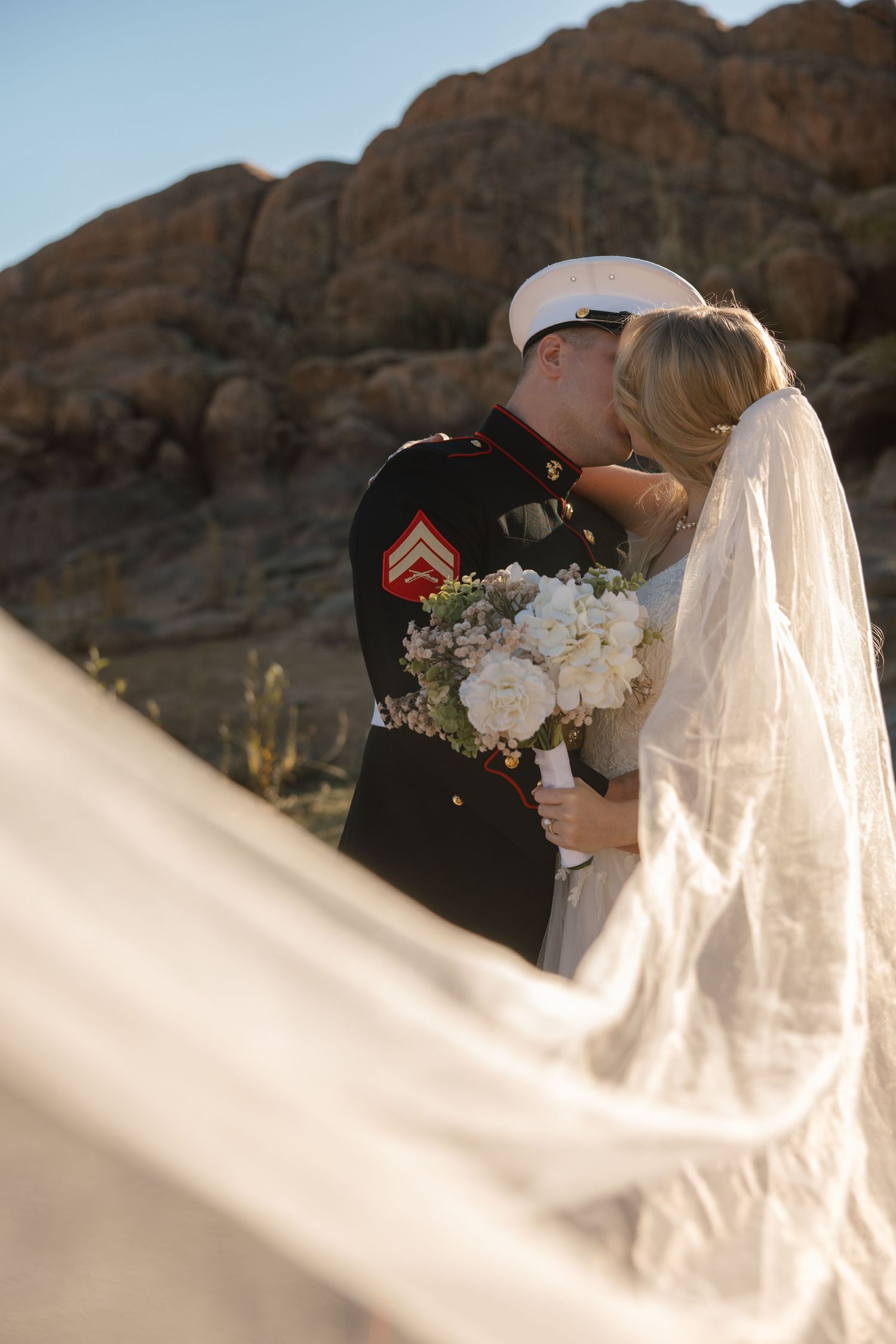 Marine in dress uniform kissing bride with flowing veil against rocky desert landscape.