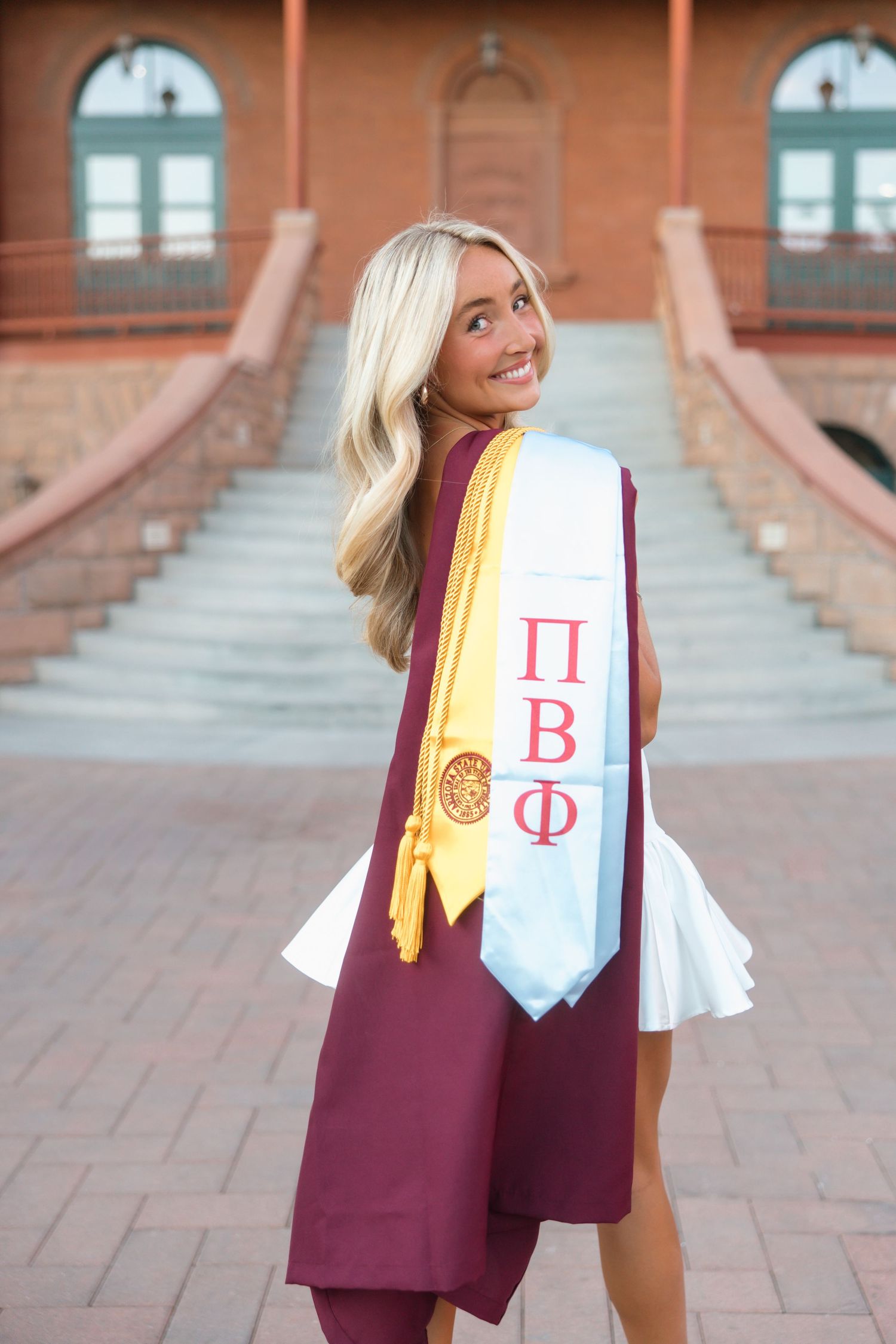 Smiling blonde woman in maroon graduation gown with Pi Beta Phi sorority sash posing outdoors on brick steps.