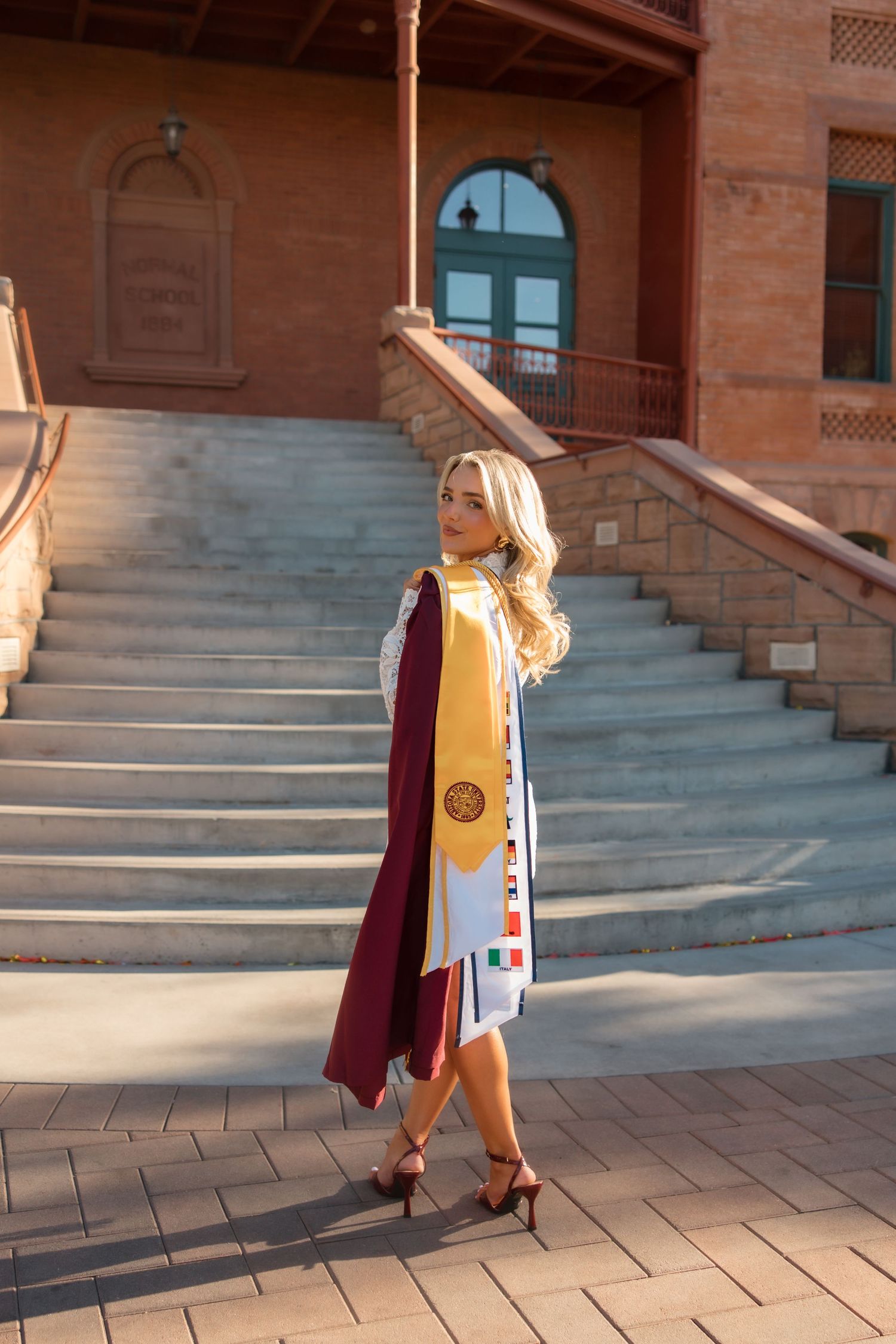 Blonde woman in maroon graduation gown with gold and white stoles poses on campus steps in heels.