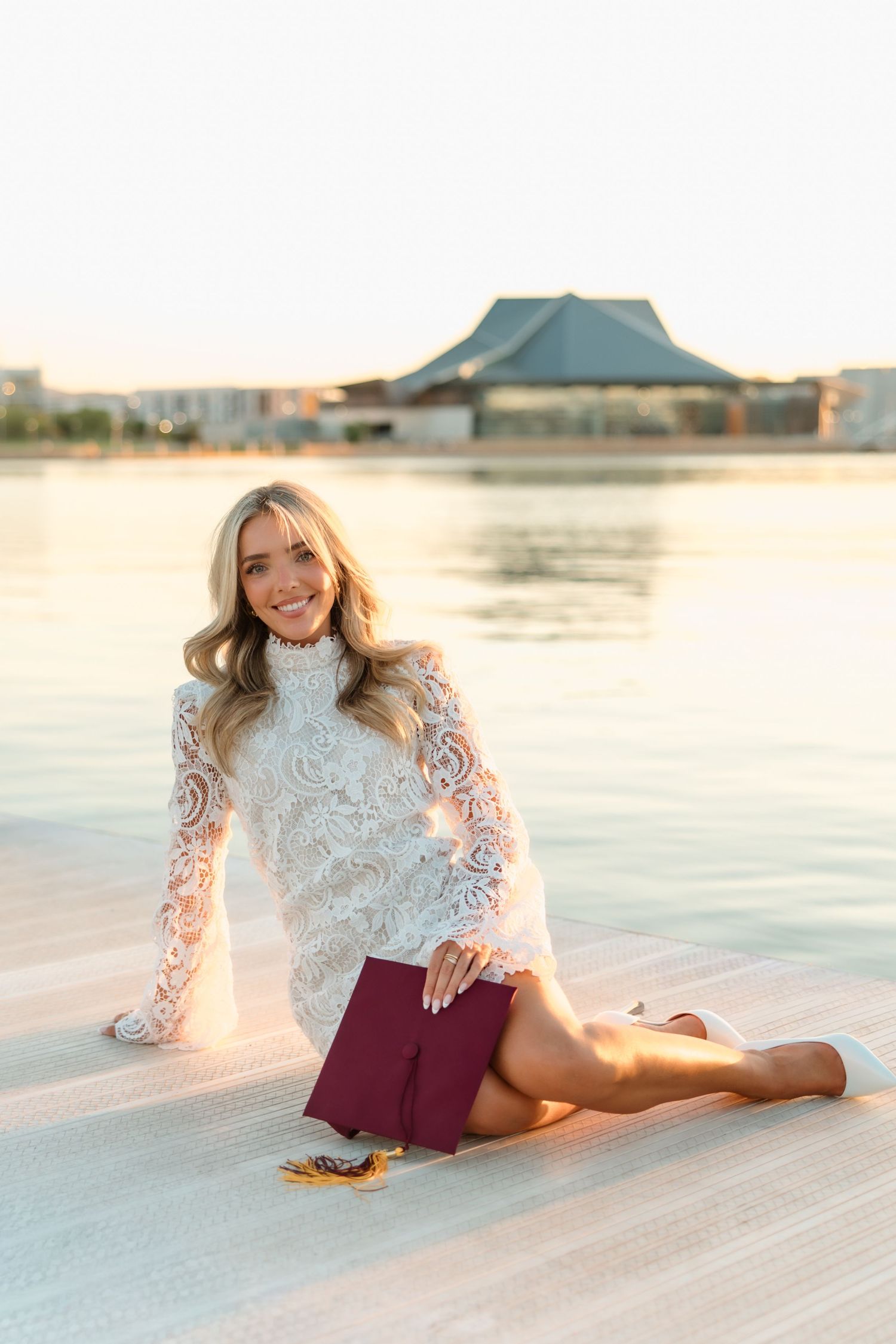 Blonde graduate in white lace top and maroon skirt sitting poolside at golden hour with diploma.