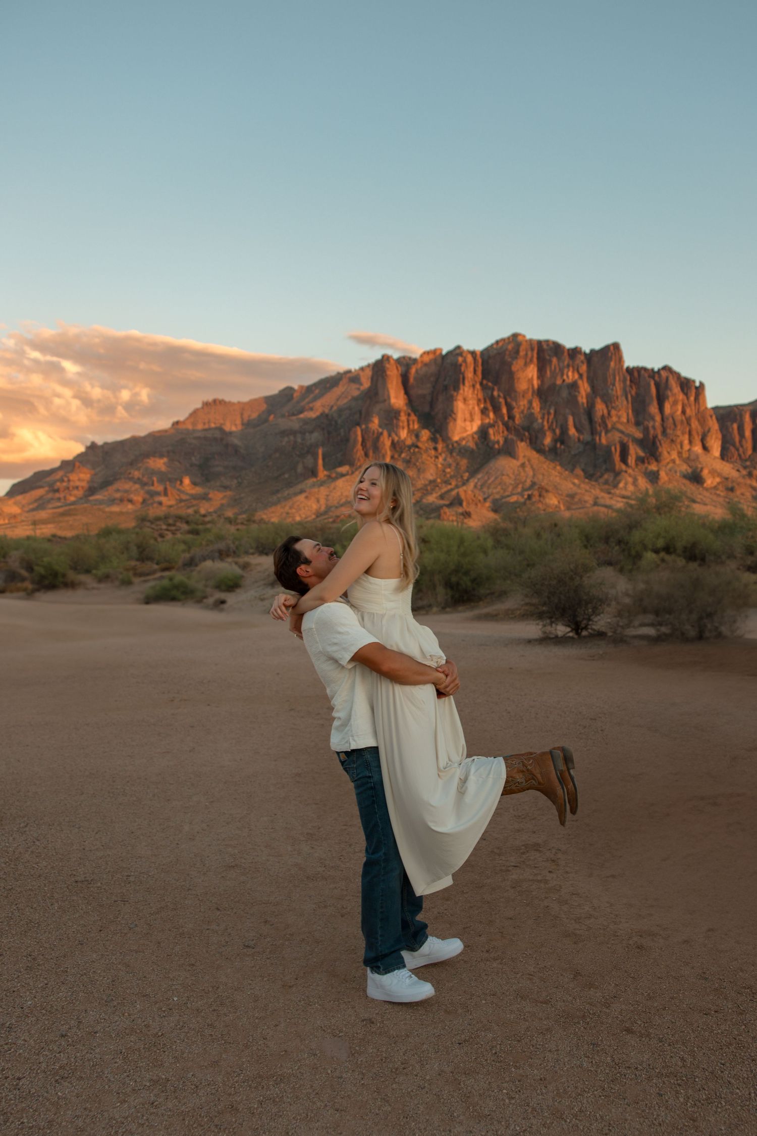 Couple embracing at sunset in Arizona desert with Superstition Mountains glowing in golden light behind them.