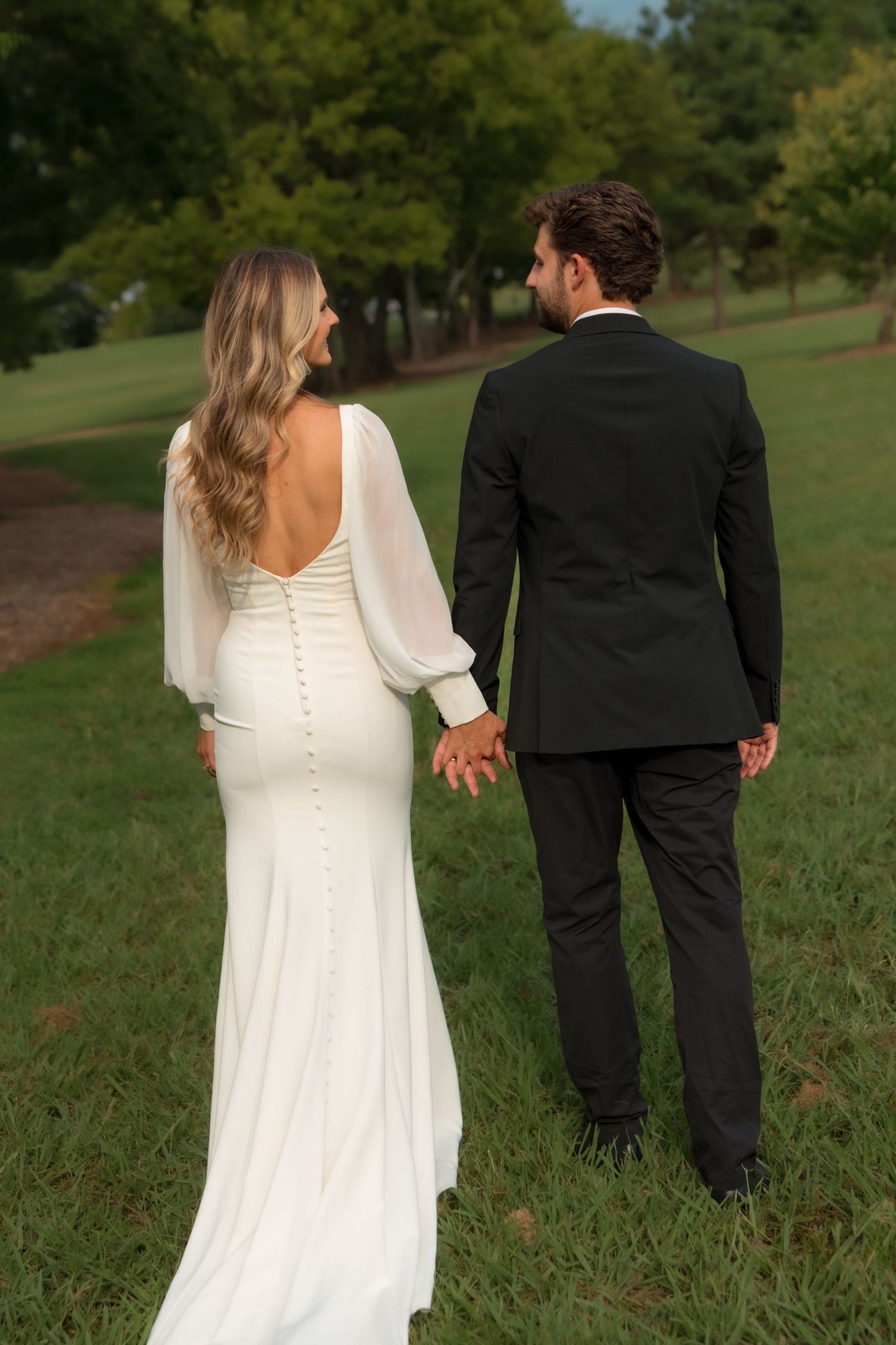 Bride and groom holding hands walking away in lush green park, wedding attire from behind.