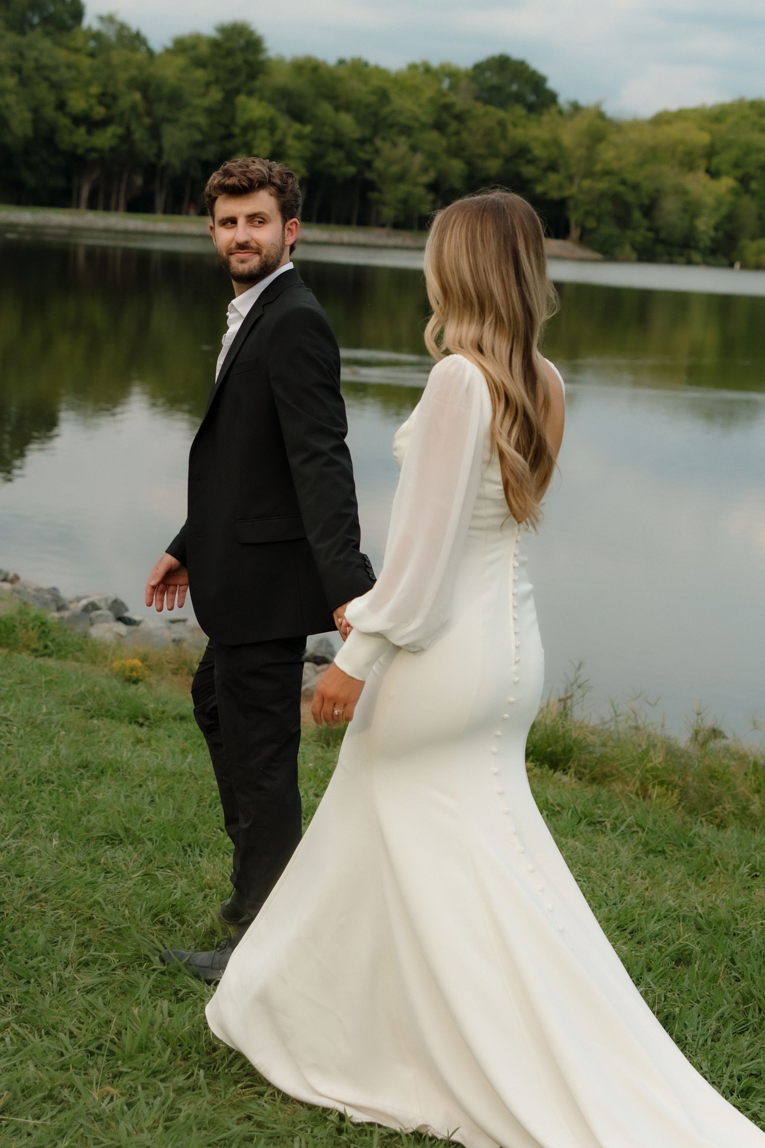 Groom in black suit leads bride in white gown along a scenic lakeside path, glancing back.
