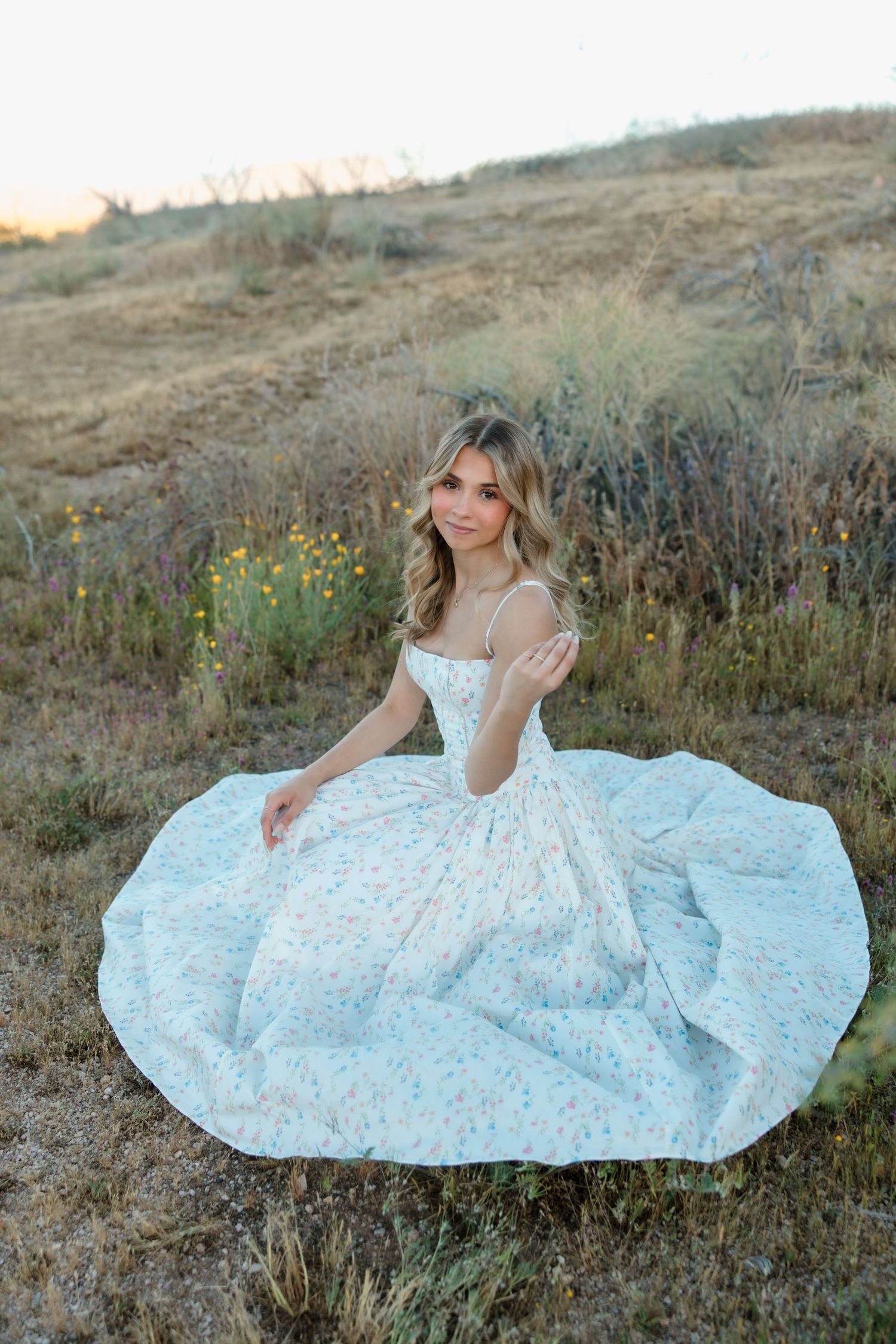 Young woman in a floral ballgown sits gracefully in a golden meadow at sunset.