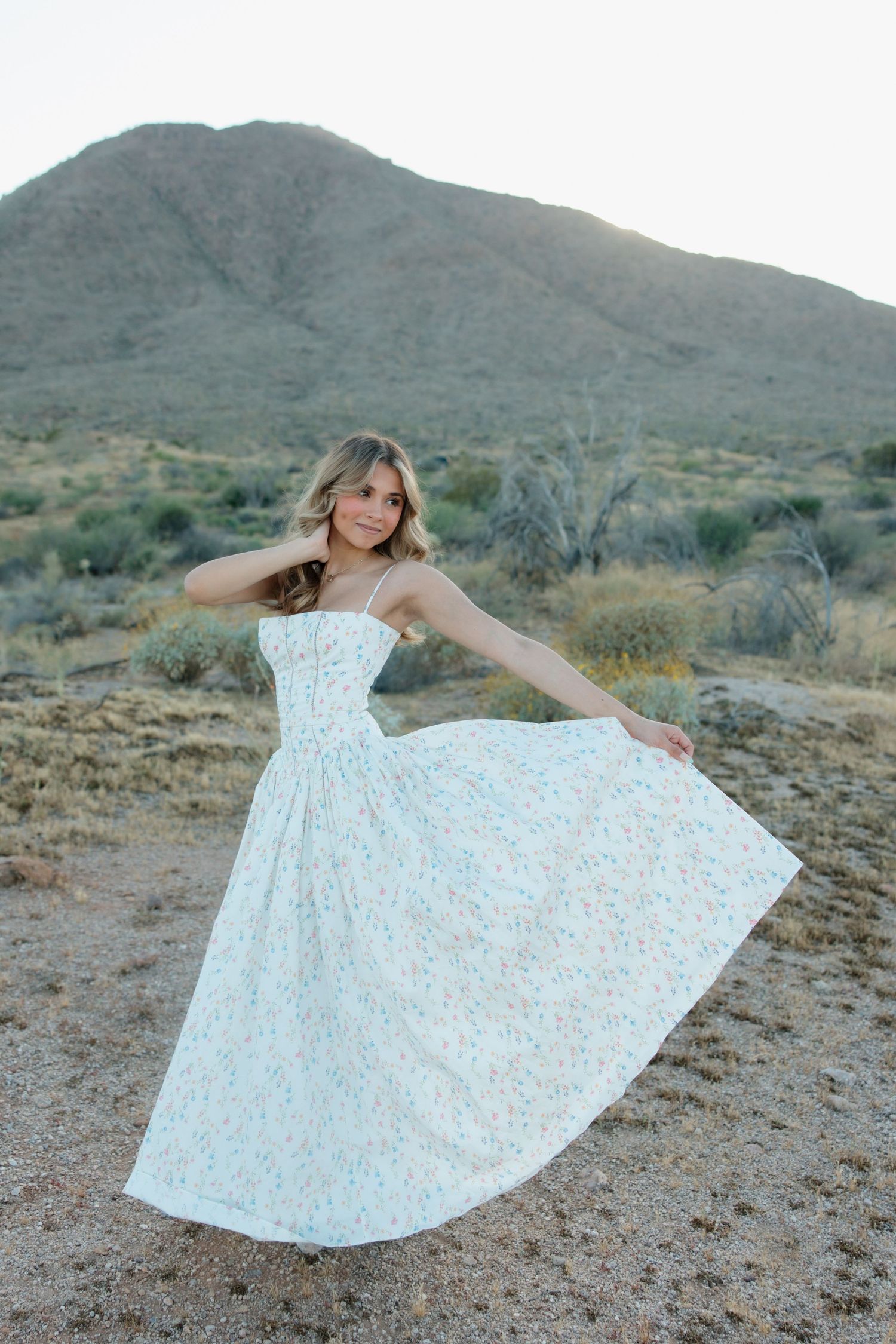 Woman in white floral midi dress twirling in desert landscape with mountain backdrop.