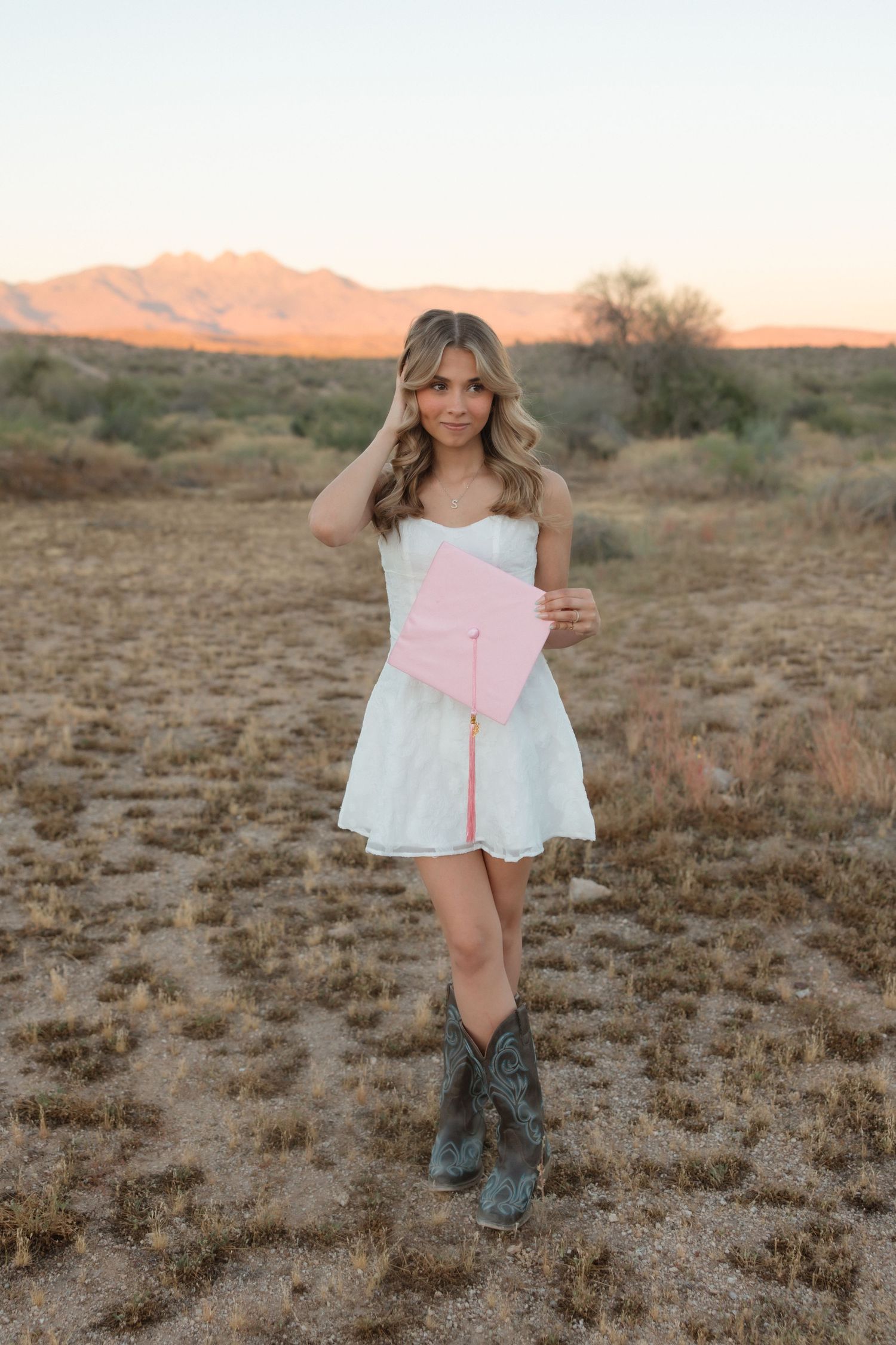 Young woman in white dress and cowboy boots holding pink graduation cap in desert landscape at sunset.