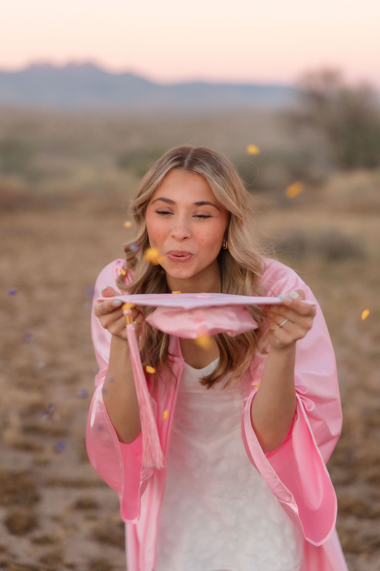 Young woman in pink robe blowing confetti from a plate outdoors in a desert landscape at sunset.