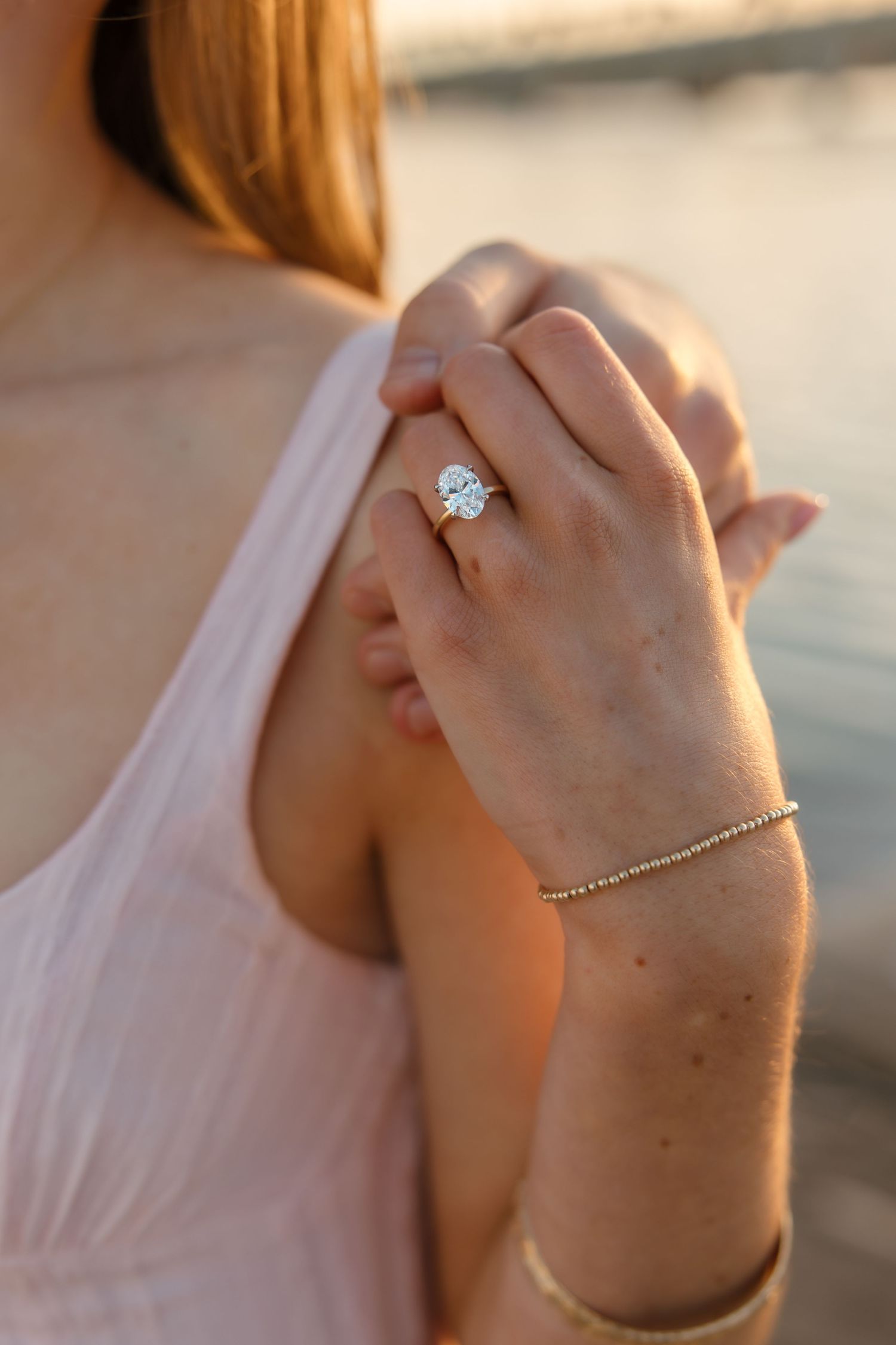 Close-up of a woman's hand showing a stunning oval diamond engagement ring with a gold bracelet at sunset.