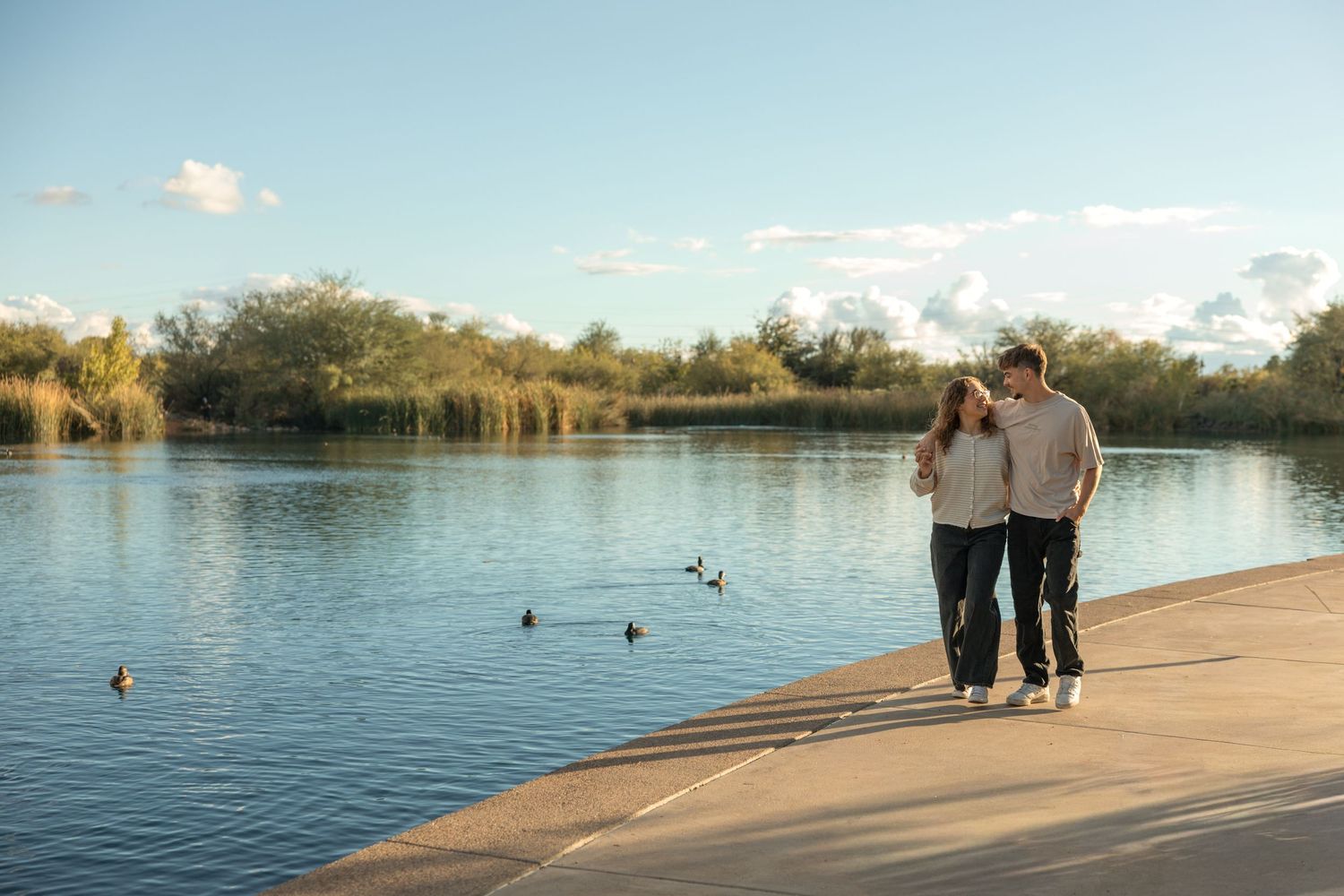 Happy couple walks hand in hand along scenic lakeside path with blue sky and lush greenery background.