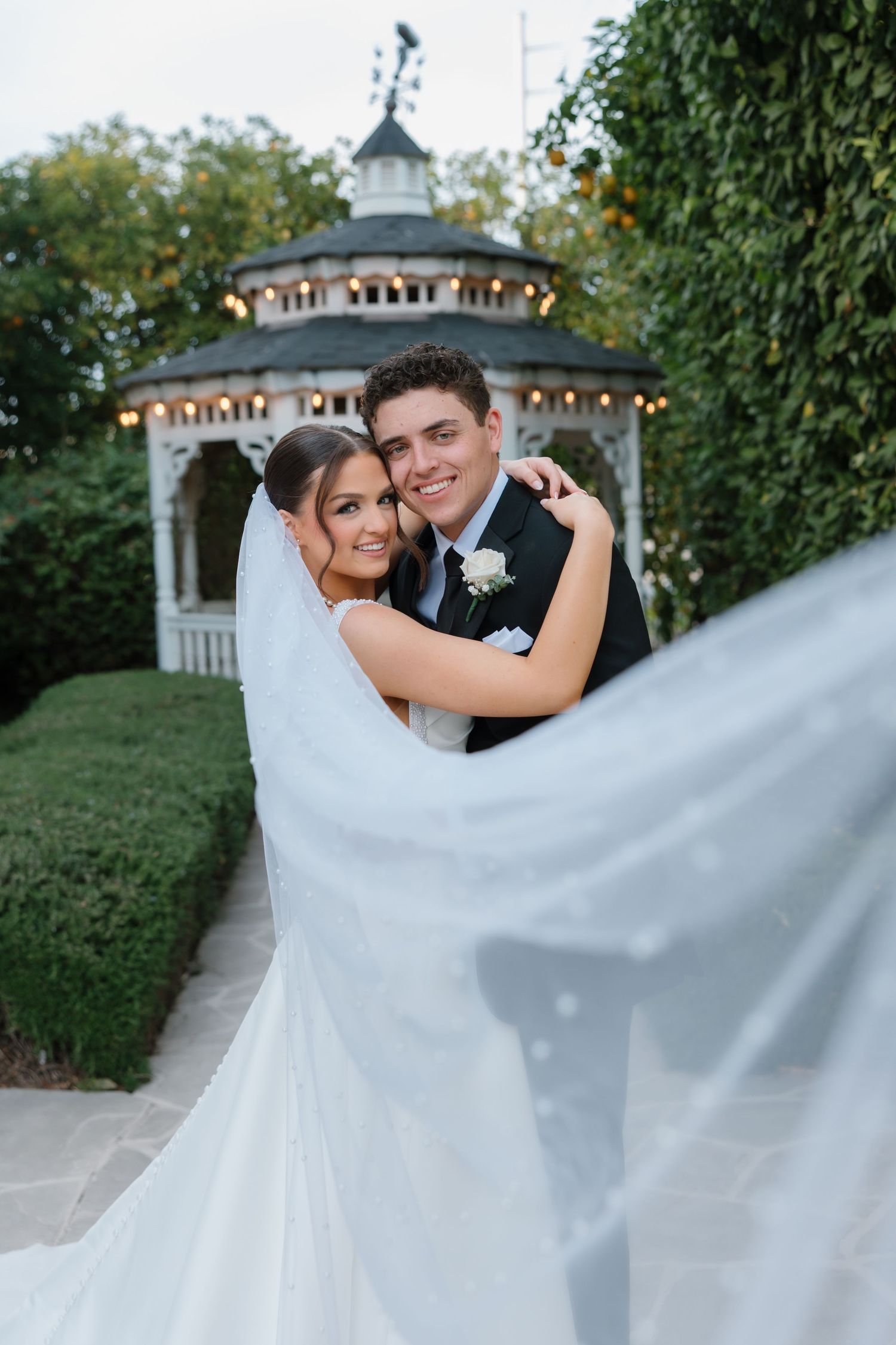 Smiling bride and groom embracing near a lit gazebo with the wedding veil flowing dramatically.