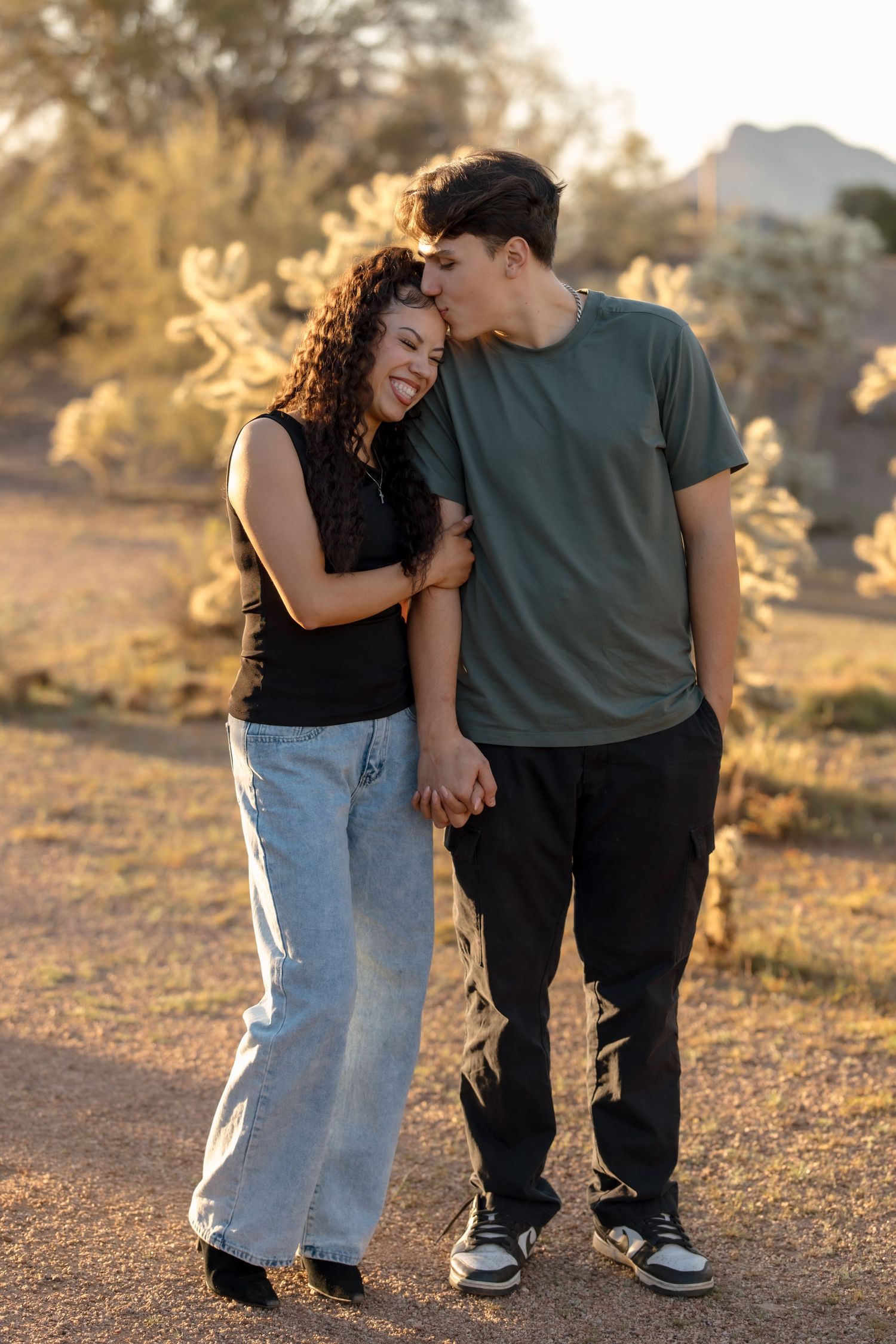 A young couple holds hands and shares a tender kiss on the forehead in a desert landscape at sunset.