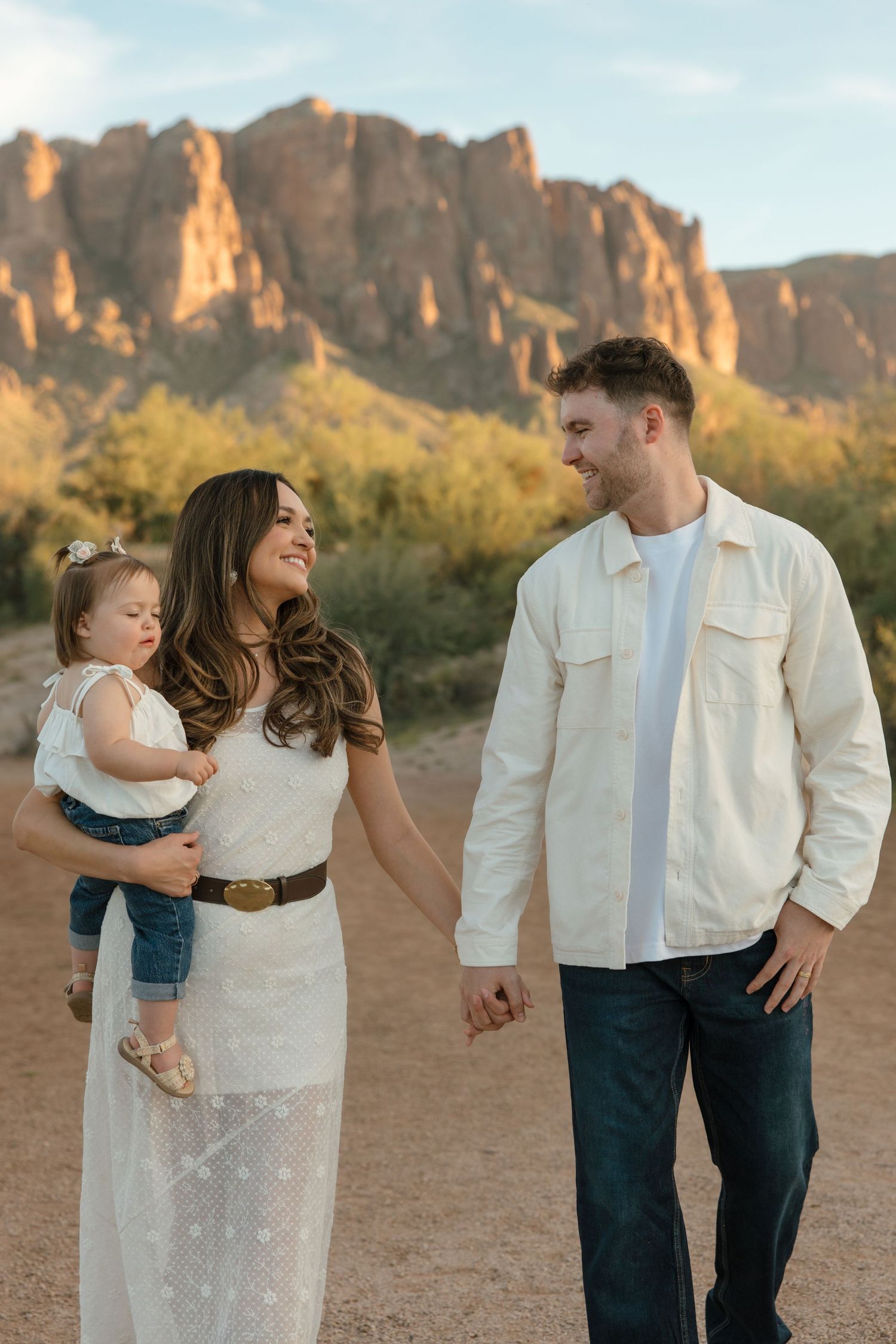 Smiling couple holding toddler daughter walking in Arizona desert with stunning mountain backdrop.