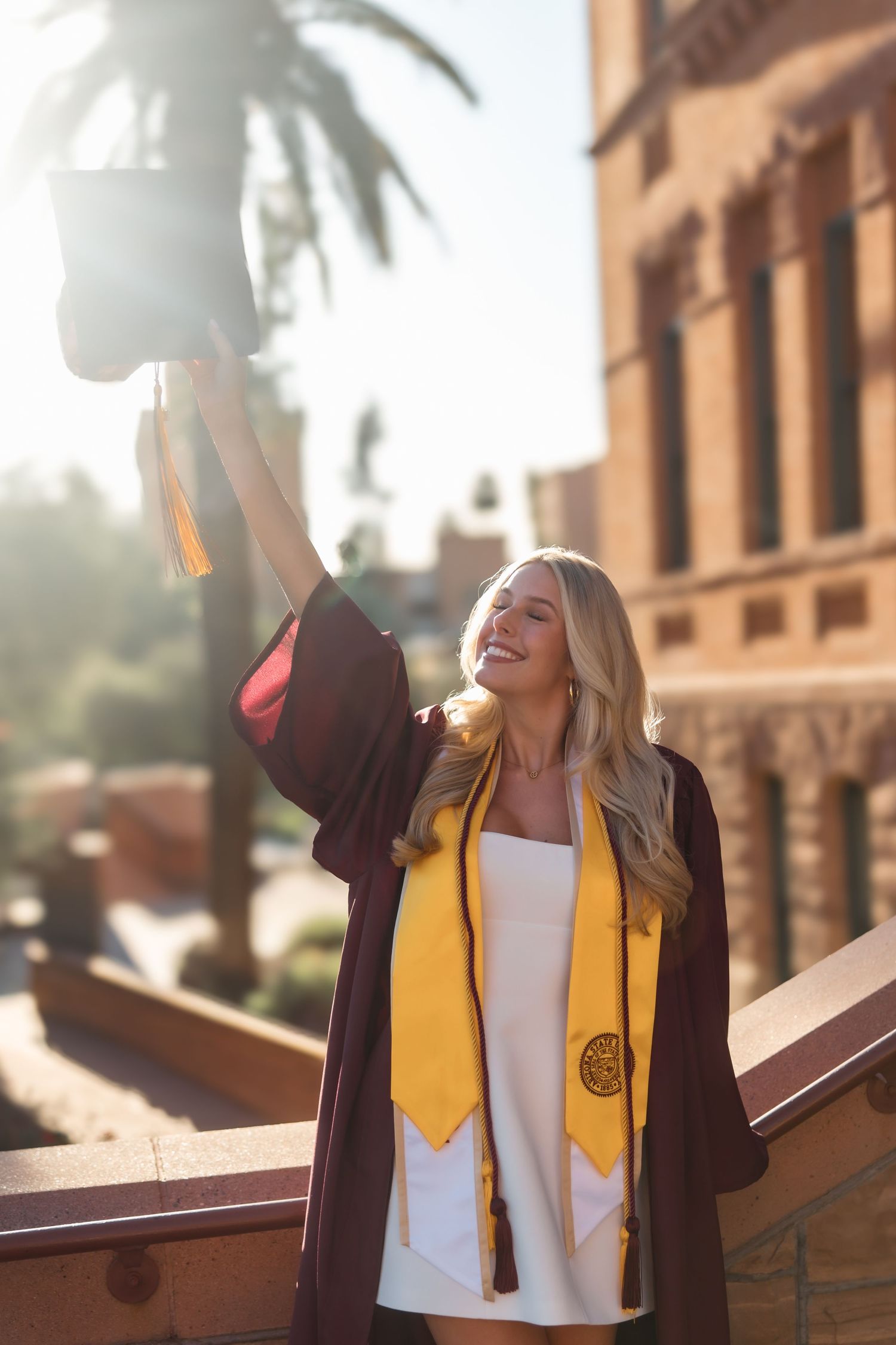 Smiling graduate in maroon gown raises cap triumphantly outdoors near palm trees and brick building.