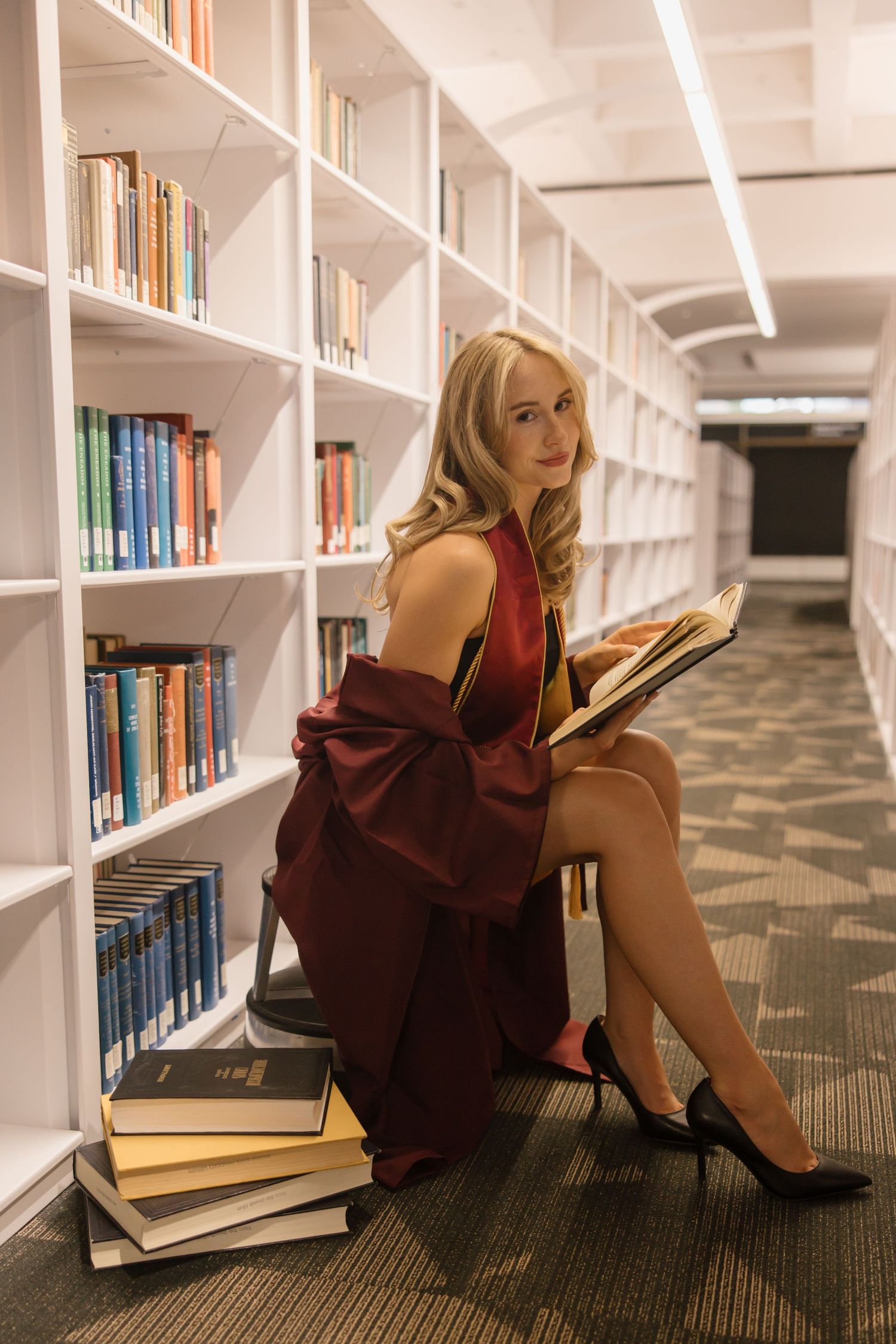 Elegant blonde woman in a burgundy dress reading a book while sitting on a stool in a modern library.