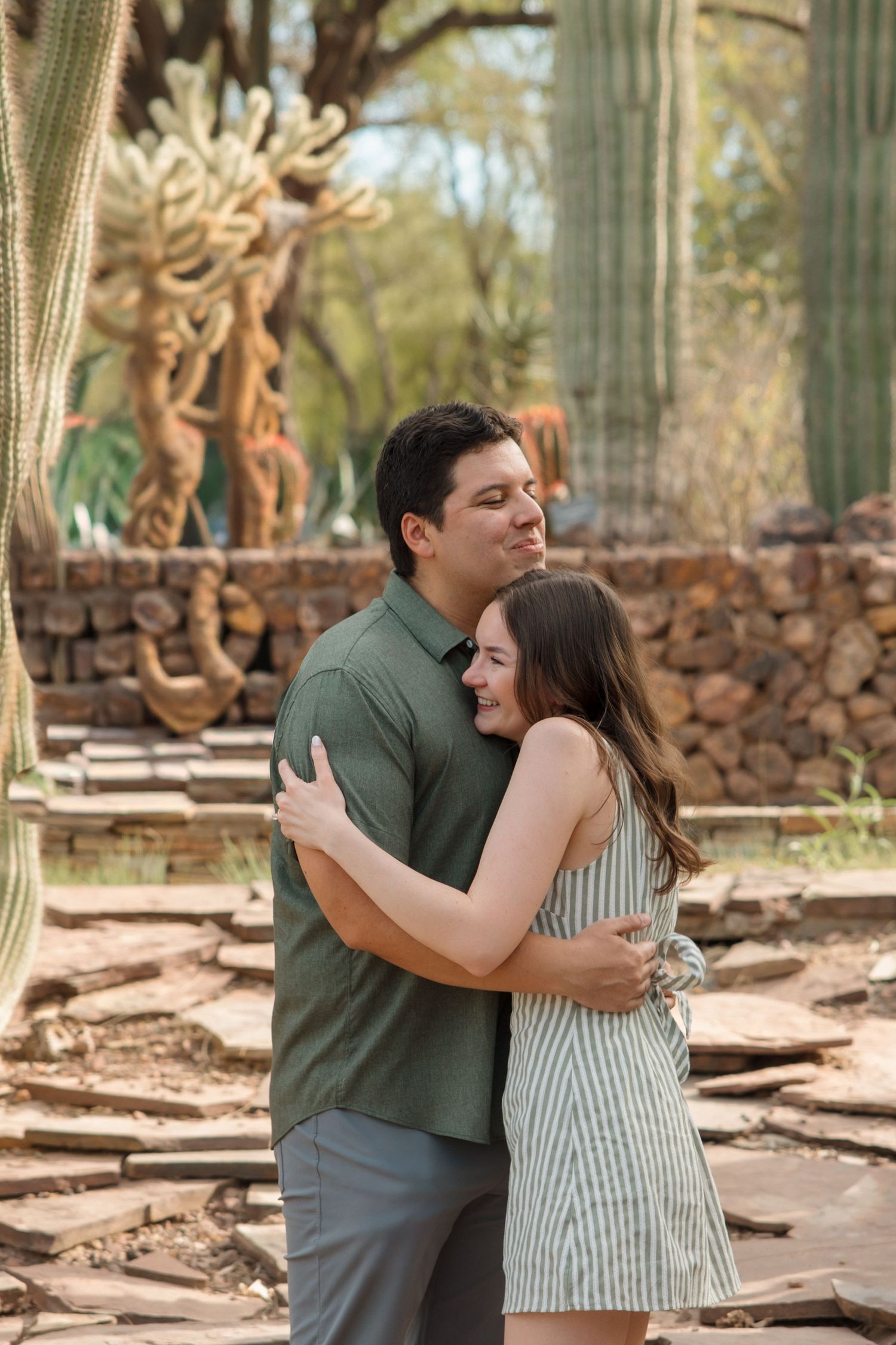 A couple embraces and smiles in a desert garden surrounded by tall saguaro cacti and stone walls.