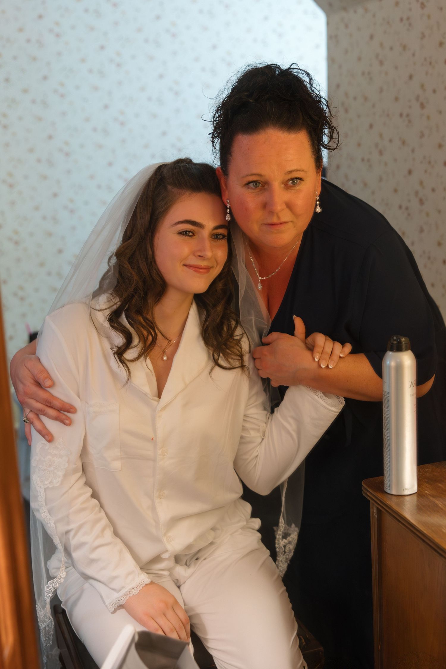 A bride in white wearing a veil sits beside an older woman in a dark top who embraces her warmly indoors.