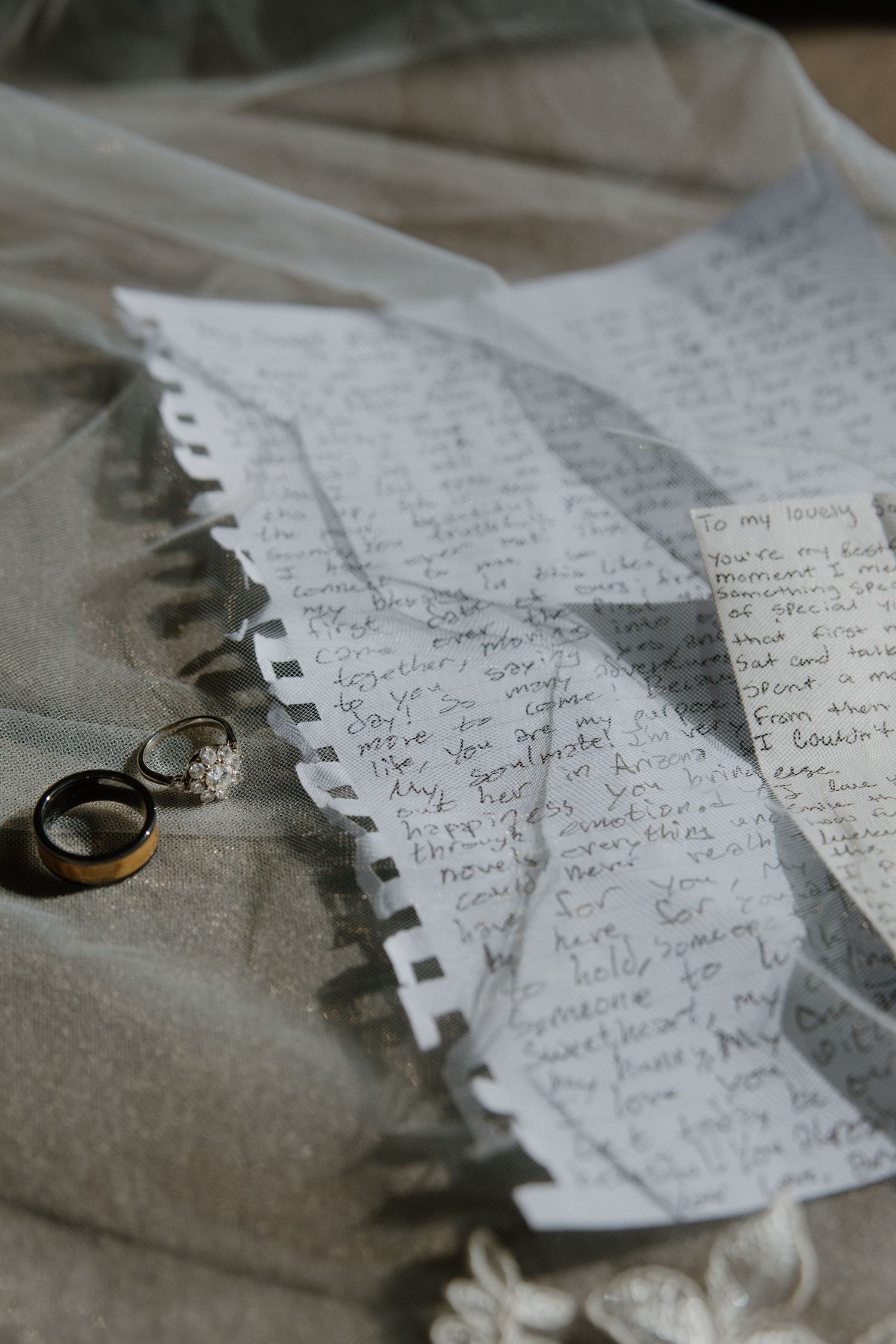 Wedding rings rest beside handwritten vows on paper draped over a lace bridal veil.