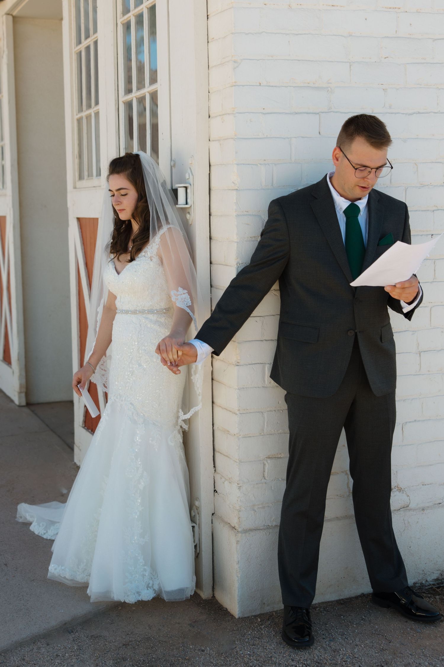 A bride and groom stand back to back reading personal wedding vows against a white brick wall.