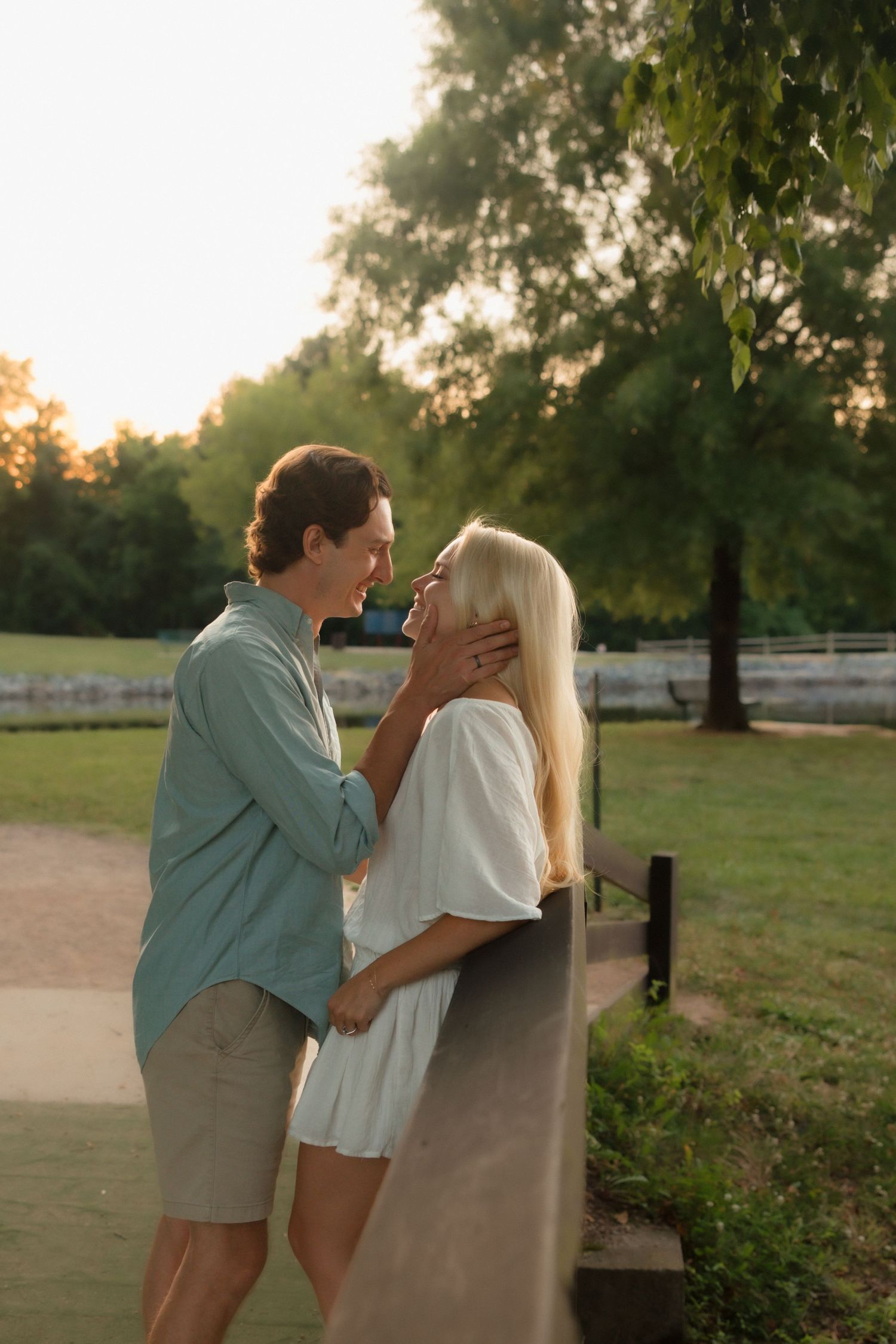 A couple shares a romantic moment leaning against a fence at golden hour in a green park.