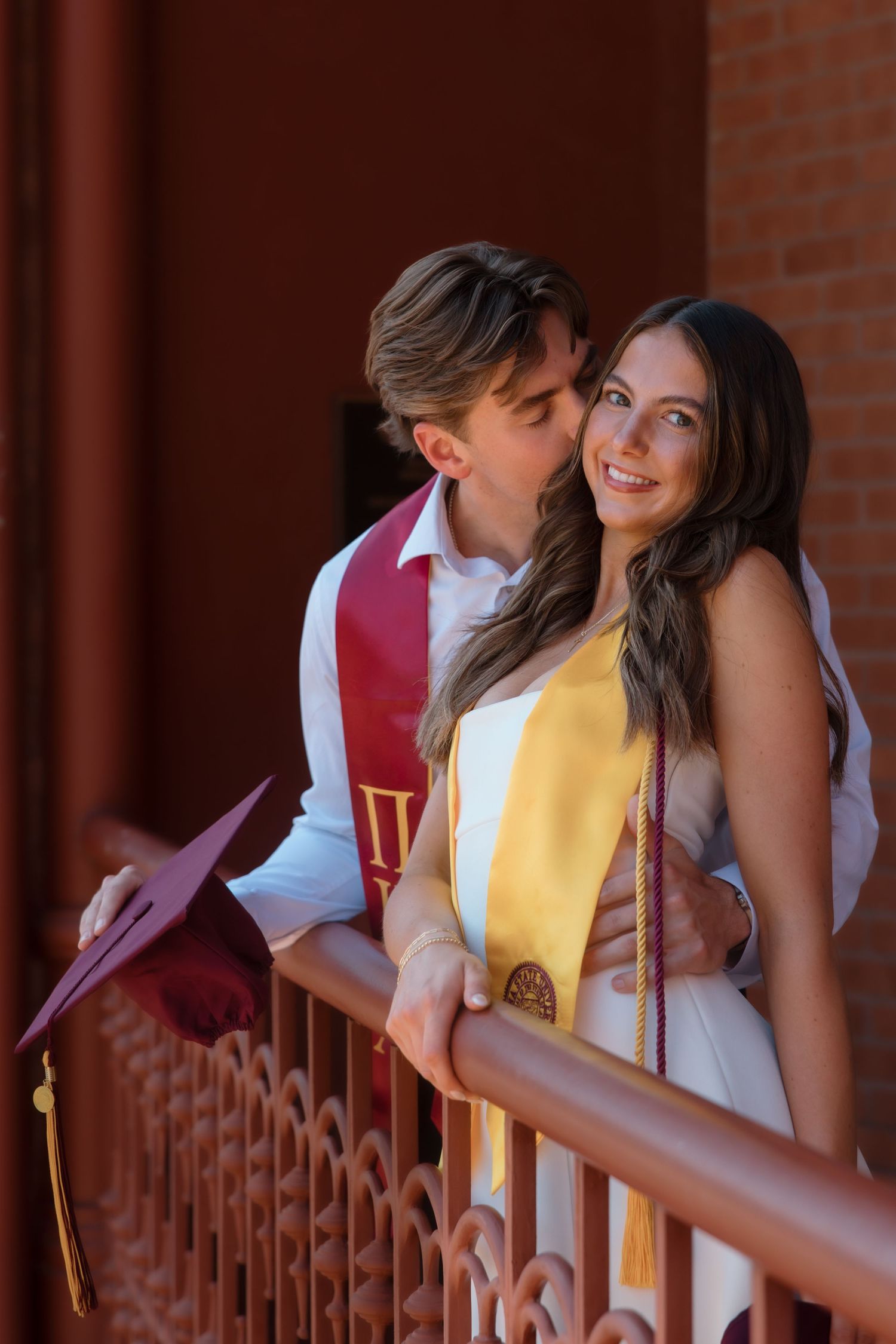 Graduate kisses woman in white dress on decorated campus balcony during graduation celebration.