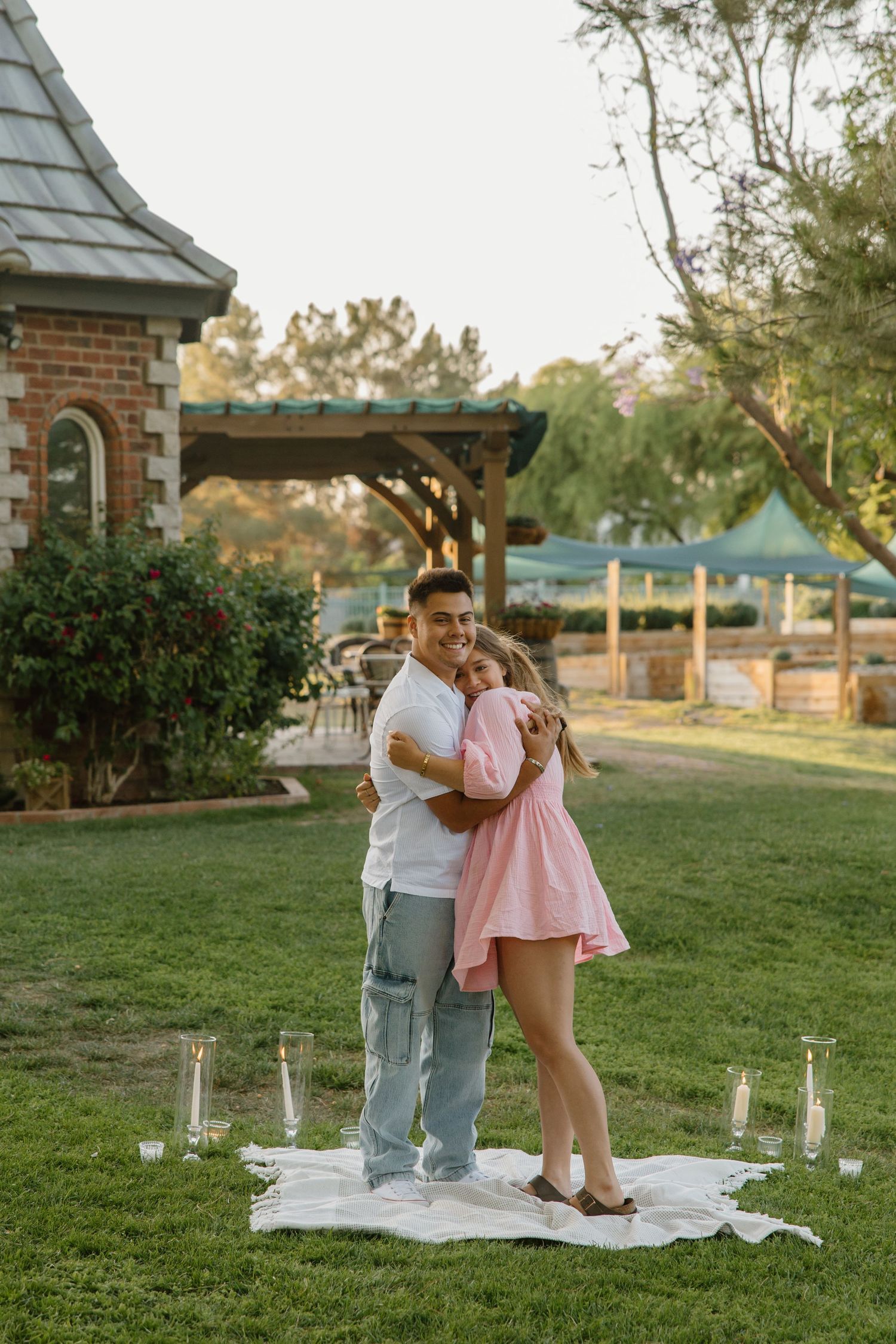 Couple embracing on a white blanket surrounded by candles in a lush garden at golden hour.