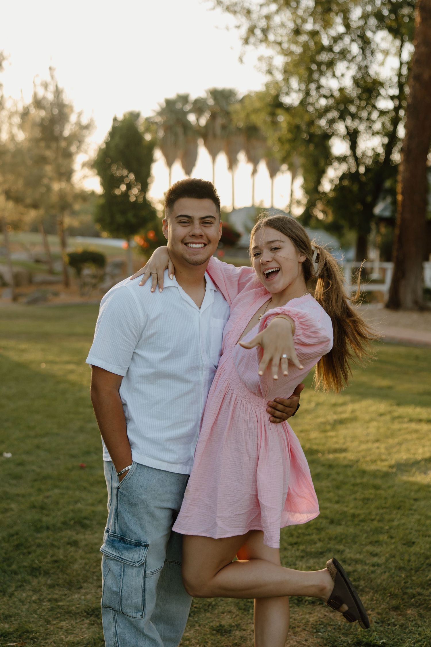Happy couple embracing outdoors at sunset, woman showing engagement ring while lifting leg in pink dress.