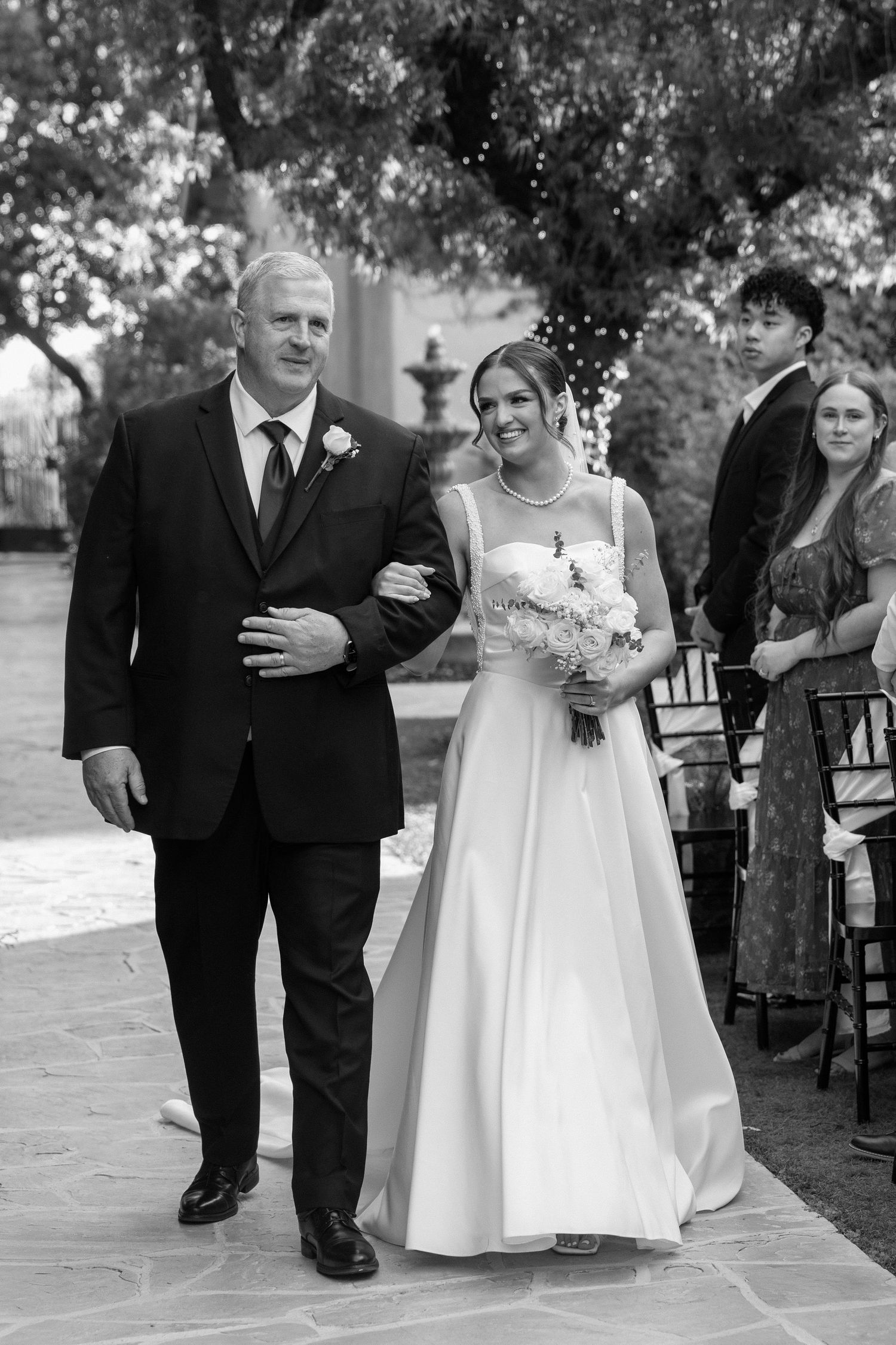 Black and white photo of father walking bride down outdoor wedding ceremony aisle.