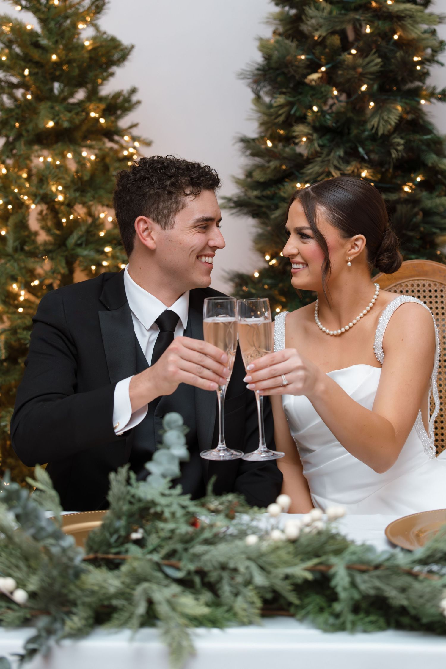 Happy bride and groom clinking champagne glasses at wedding reception with decorated Christmas trees in background.