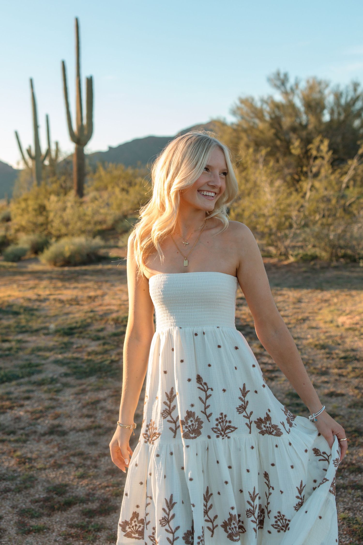 Joyful blonde woman twirling white floral dress in golden hour Arizona desert setting.