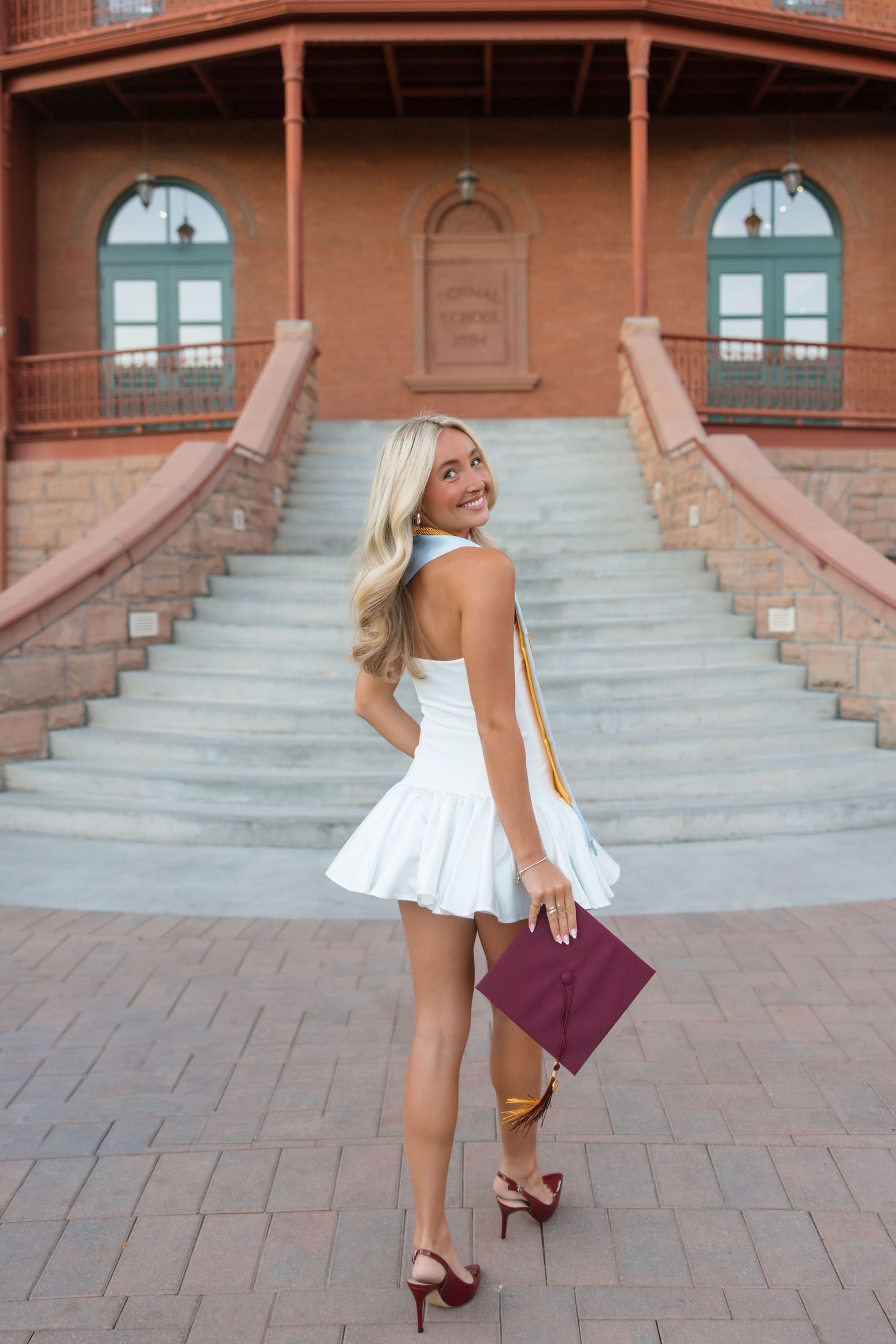 Smiling blonde woman in white mini dress holding maroon graduation cap on brick steps.