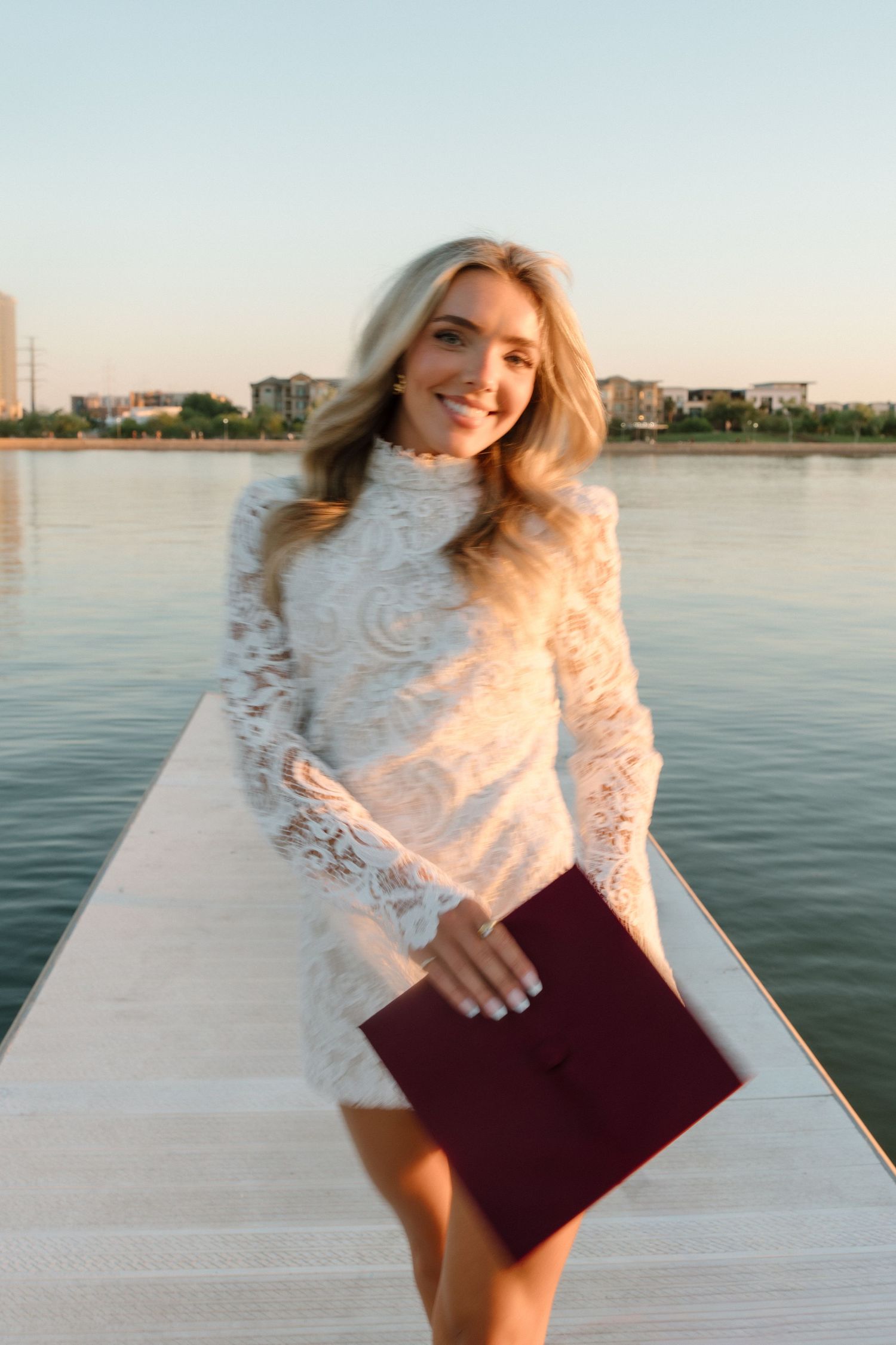 Smiling blonde woman in white lace dress holds maroon diploma folder on waterfront pier.