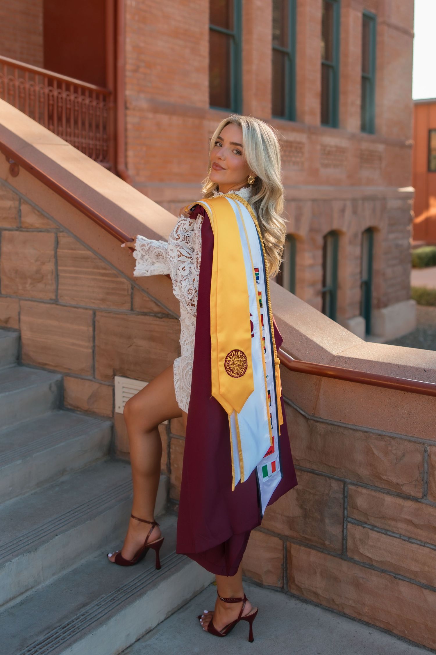 Graduate in maroon gown with gold honor stole poses on brick university staircase wearing heels.