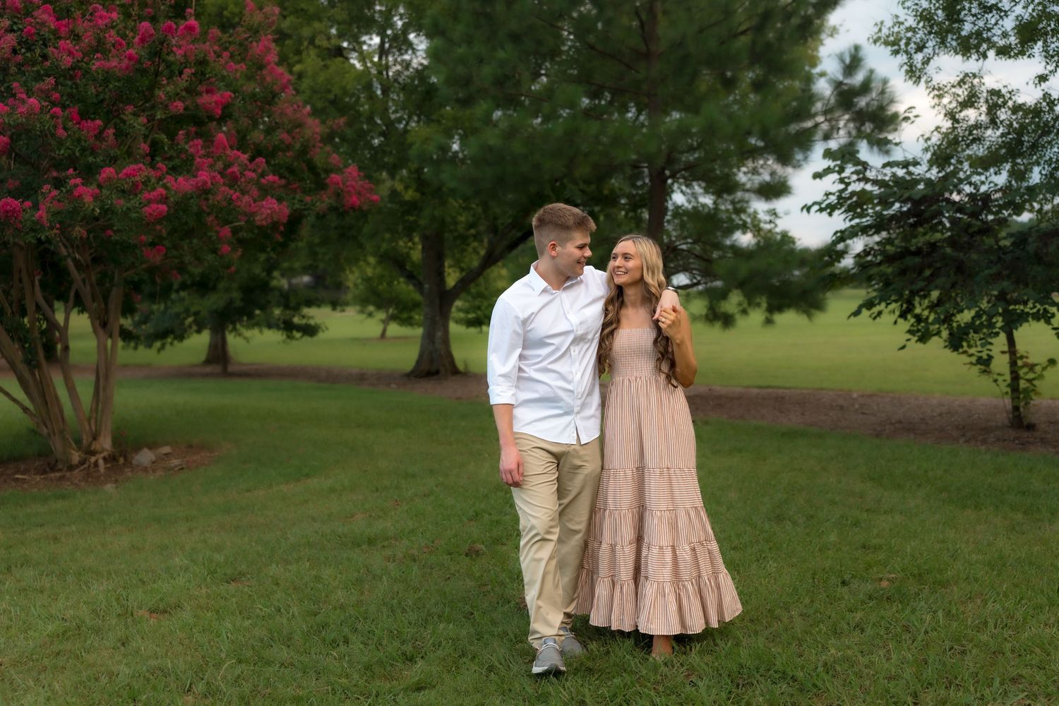 Couple walking hand in hand in a lush green park with blooming red crepe myrtle trees at dusk.