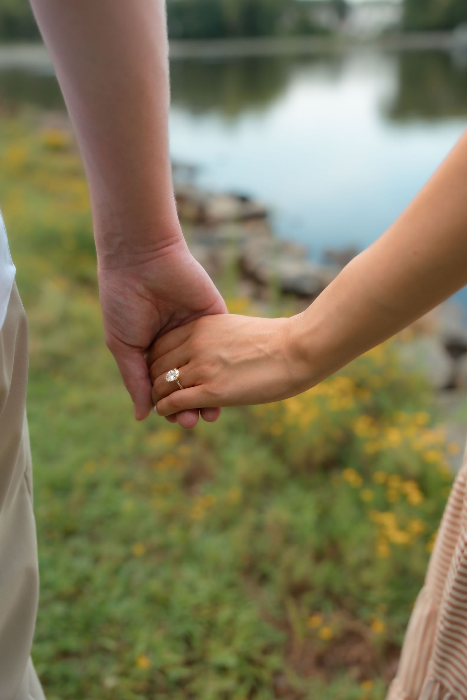 Couple holding hands near a lake with wildflowers, woman wearing an engagement ring, romantic outdoor setting.