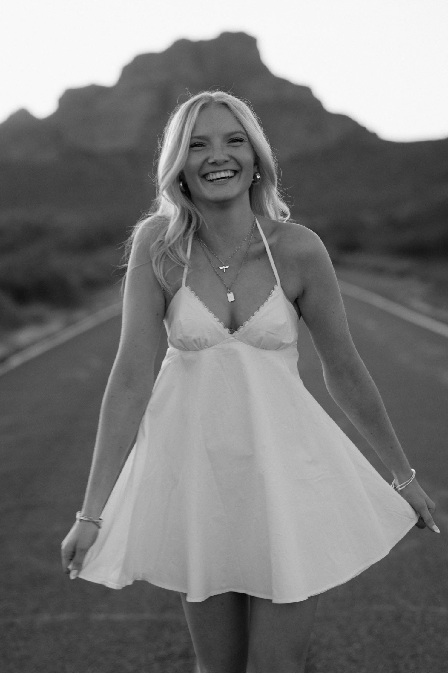 Smiling blonde woman in white halter dress standing on open road with mountain backdrop, black and white photo.