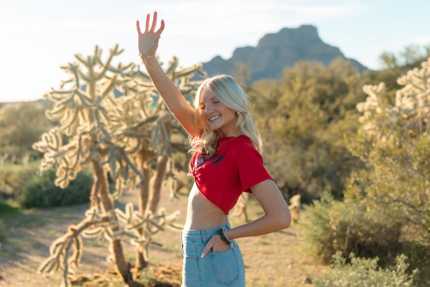 Blonde woman in red crop top and jeans waving in desert landscape with cacti and mountain backdrop.