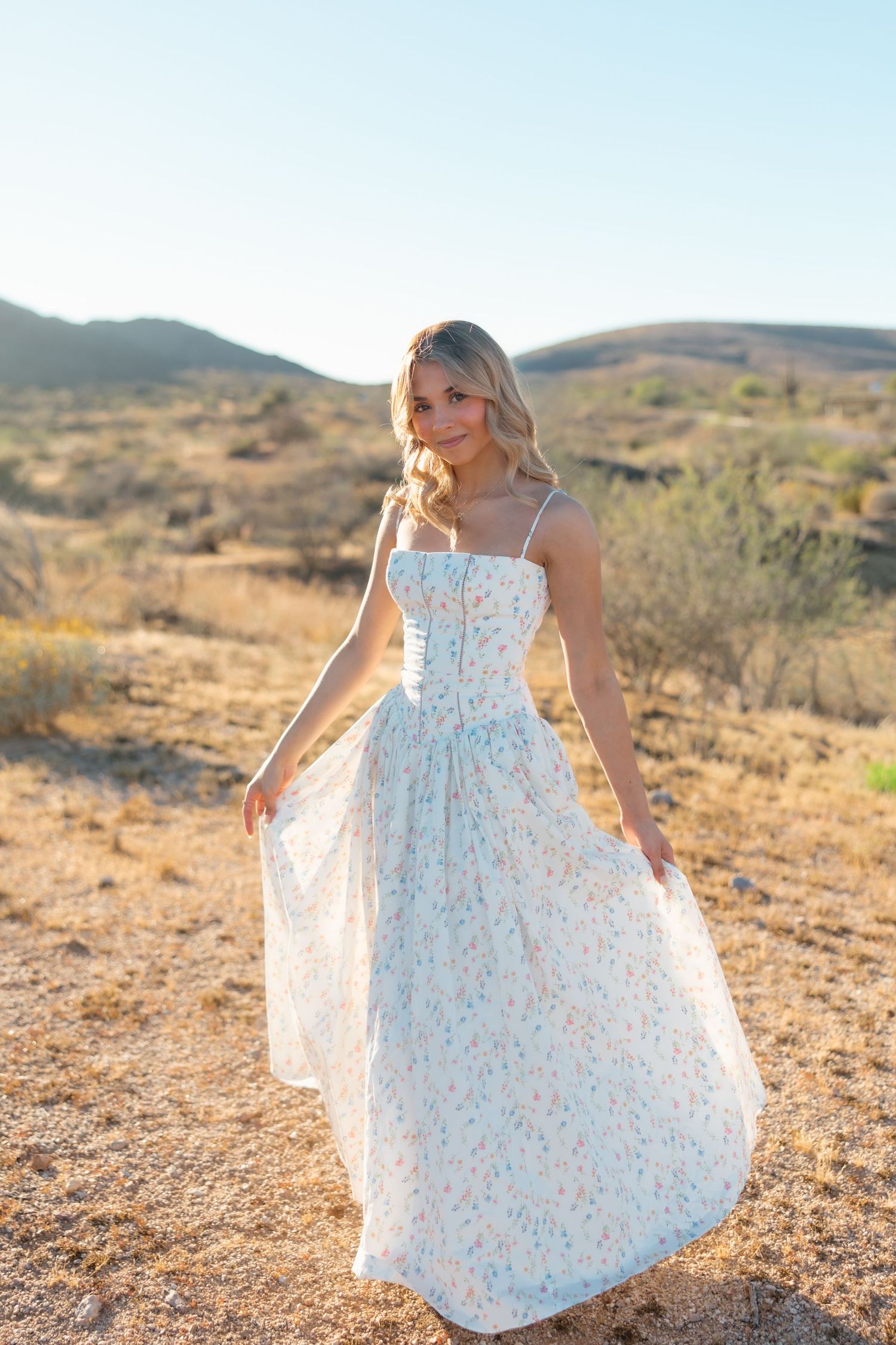 A blonde woman in a floral maxi dress poses gracefully in a desert landscape with mountains in the background.