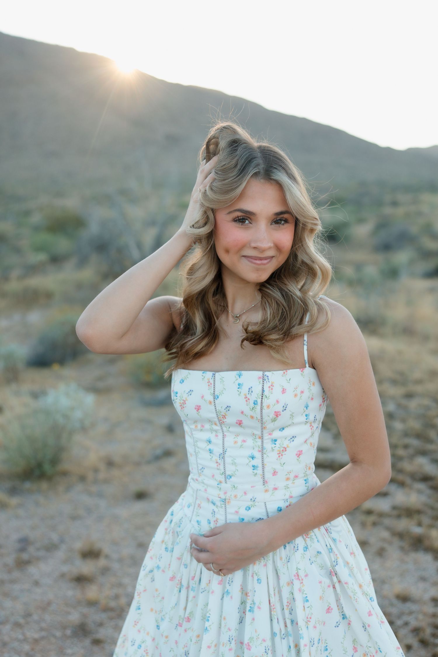 Young woman in floral sundress poses outdoors in desert landscape at golden hour sunset.